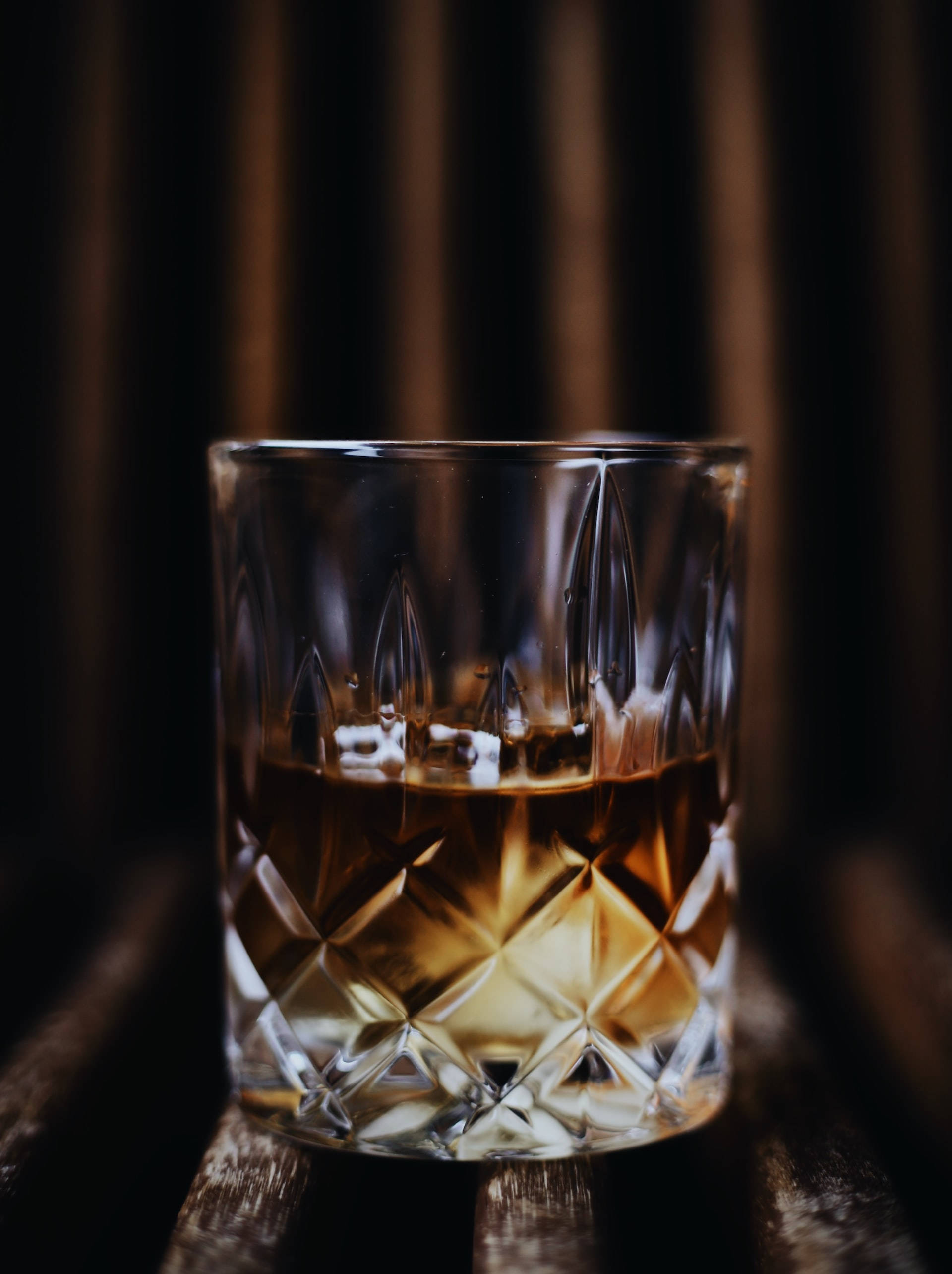alcohol 桌布 壁紙 — 一個水晶切割的威士忌杯，盛裝著琥珀色液體，放在質樸的木質表面上，背景是深色模糊的。