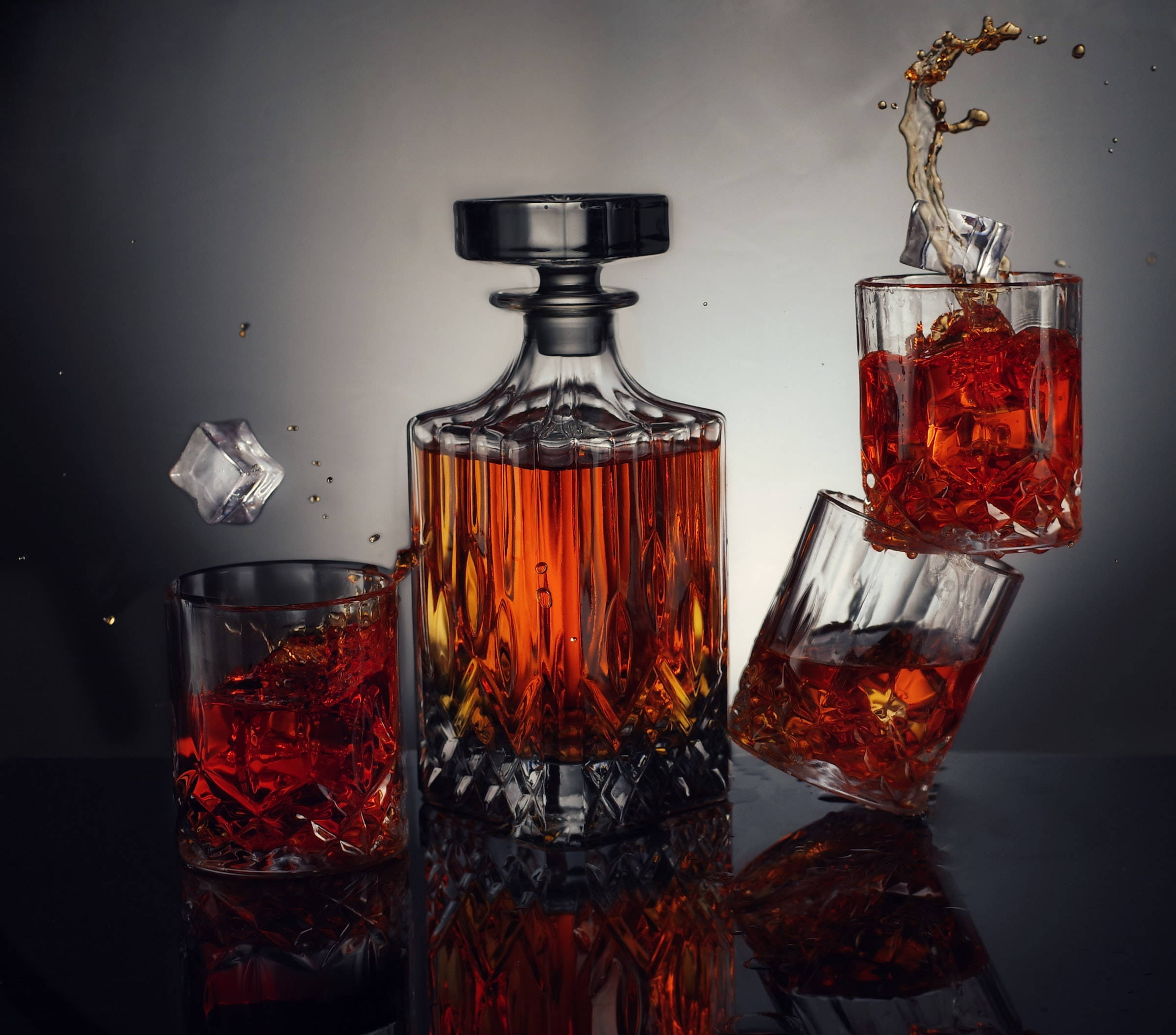 alcohol 桌布 壁紙 — 一個水晶醒酒器和玻璃杯裝滿琥珀色液體和冰塊，在深色漸變背景上捕捉動態飛濺。