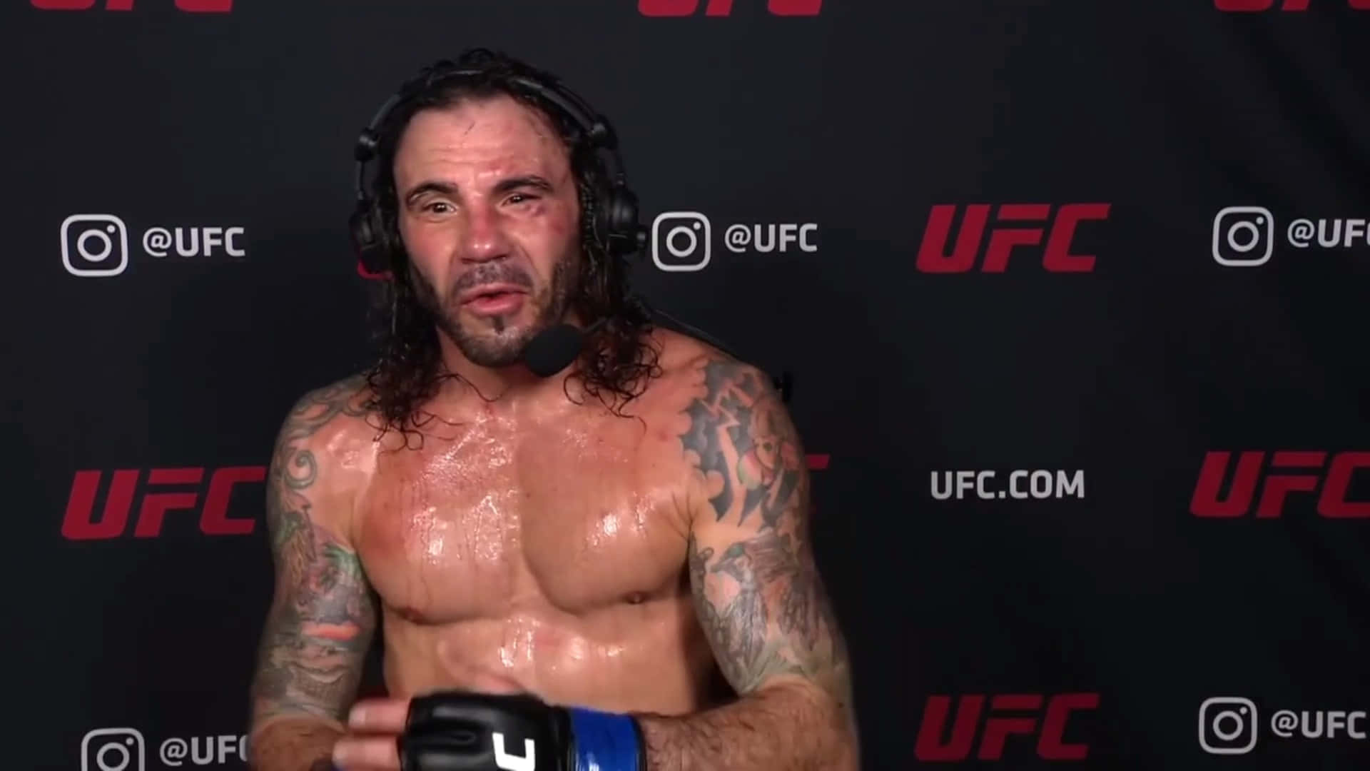 UFC 桌布 壁紙 — 一個汗流浹背、有刺青的UFC拳手戴著耳機，在品牌黑色背景前對著麥克風說話。