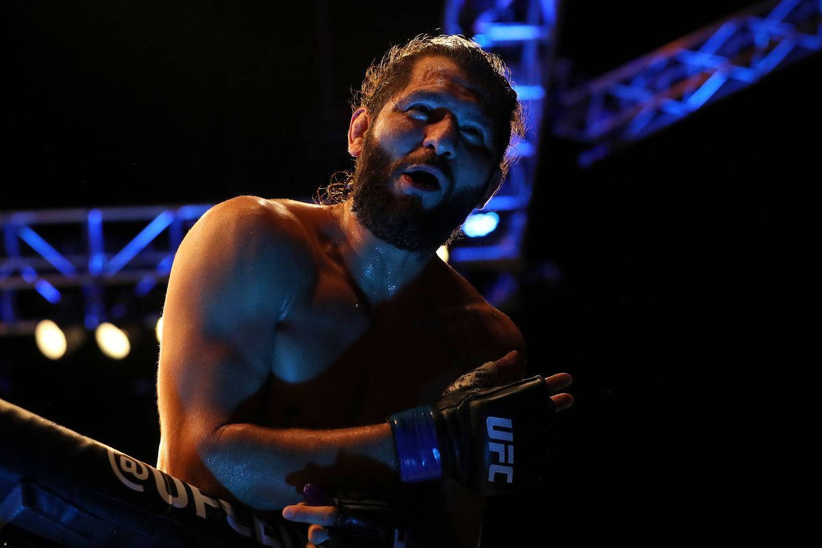 UFC 桌布 壁紙 — 一個汗流浹背、留鬍子的UFC拳手在戲劇性的藍色舞台燈光下靠在八角籠的繩索上。