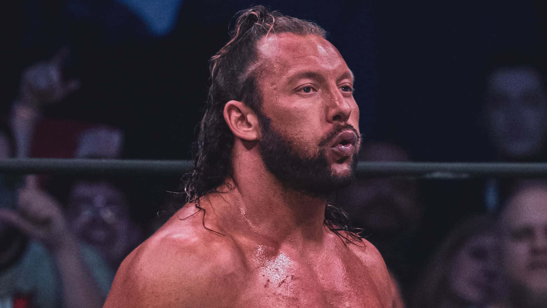 Aew 桌布 壁紙 — 一個汗流浹背、留鬍子的摔角手在明亮的場館燈光下，在擂台上專注地凝視，背後是模糊的人群。