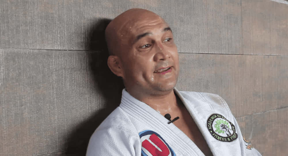 MMA 桌布 壁紙 — 一個汗流浹背的光頭男人穿著白色武術道服，靠著紋理灰色牆壁，訓練後顯得疲憊。