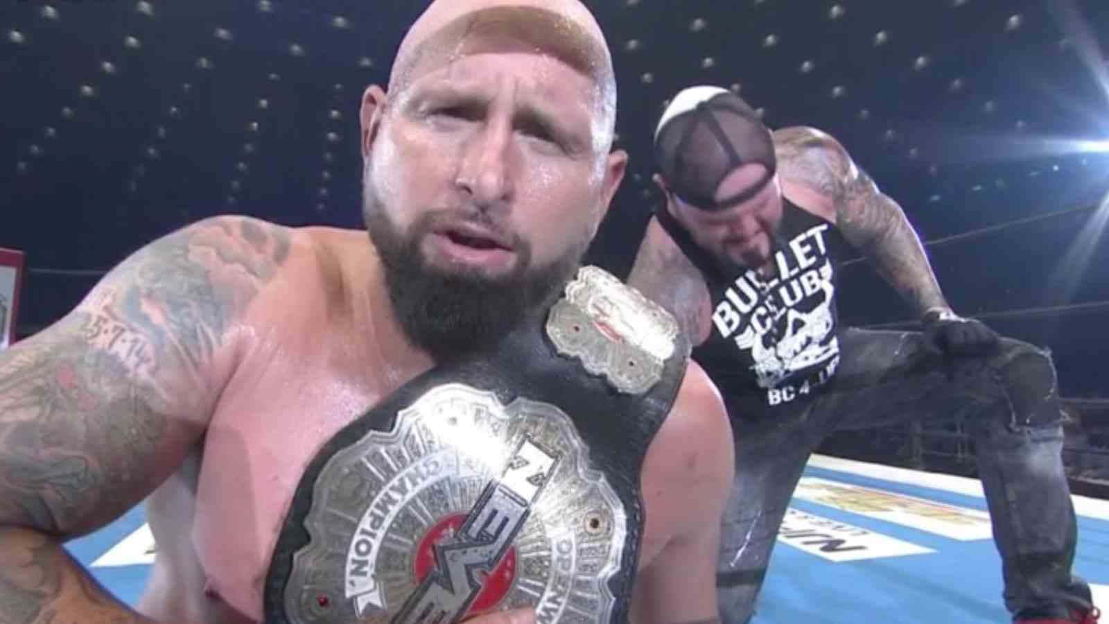 WWE 桌布 壁紙 — 一個汗流浹背的光頭鬍鬚摔角手拿著冠軍腰帶，而另一個紋身摔角手穿著Bullet Club T恤站在他身後擂台上。