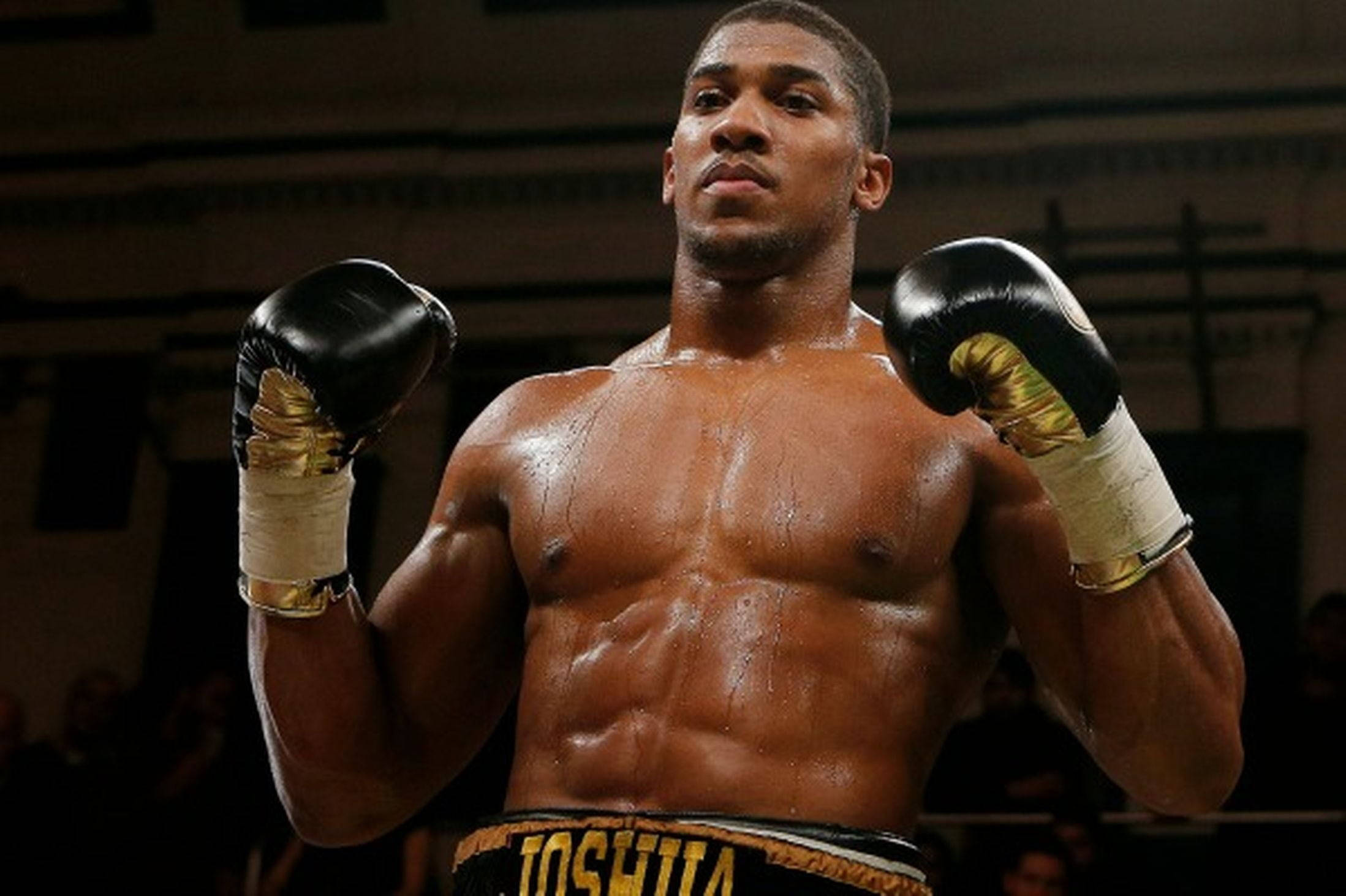 Anthony Joshua 桌布 壁紙 — 一個汗流浹背的肌肉發達拳擊手站在擂台上，戴著黑色和金色的手套，專注而強烈。