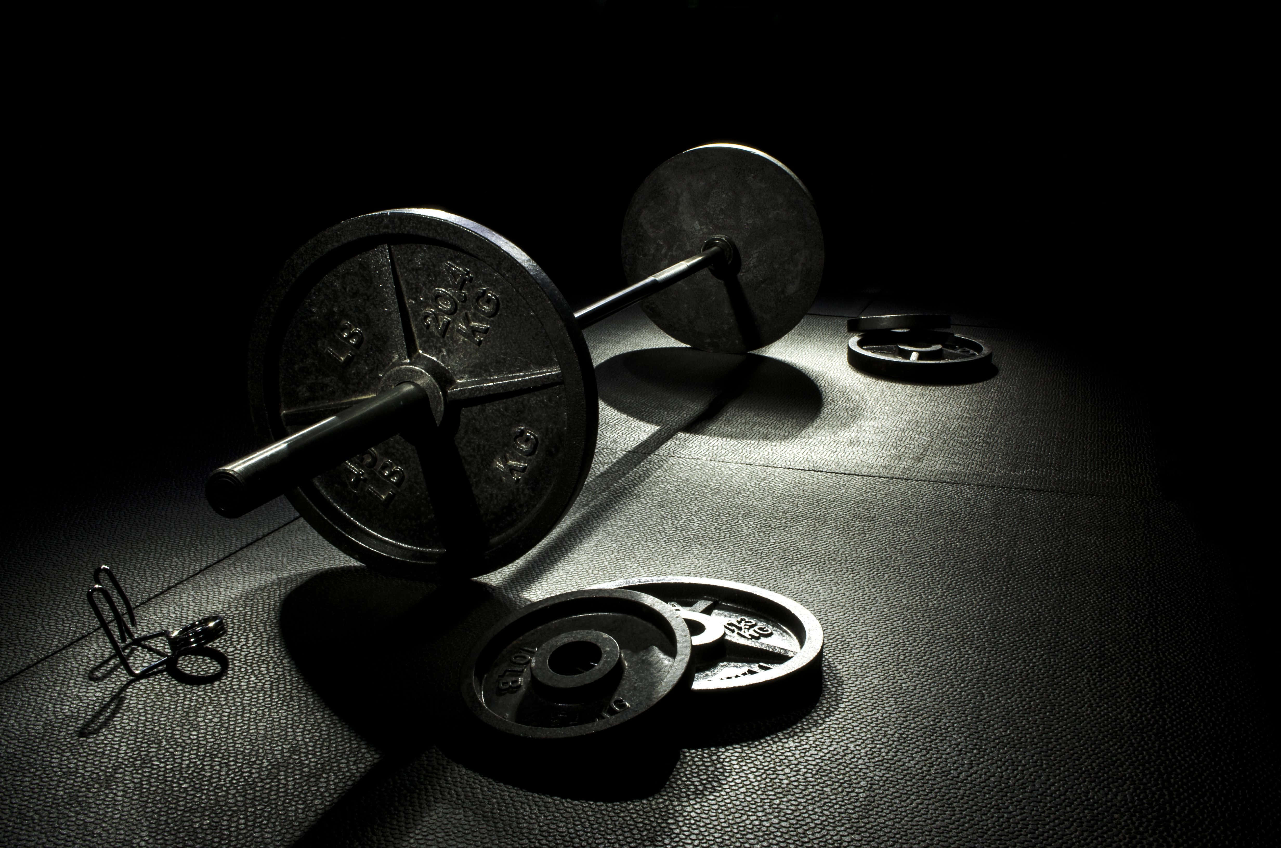 Barbell 桌布 壁紙 — 一個沉重的槓鈴和啞鈴在黑色背景的強烈聚光燈下，戲劇性地放在健身墊上。