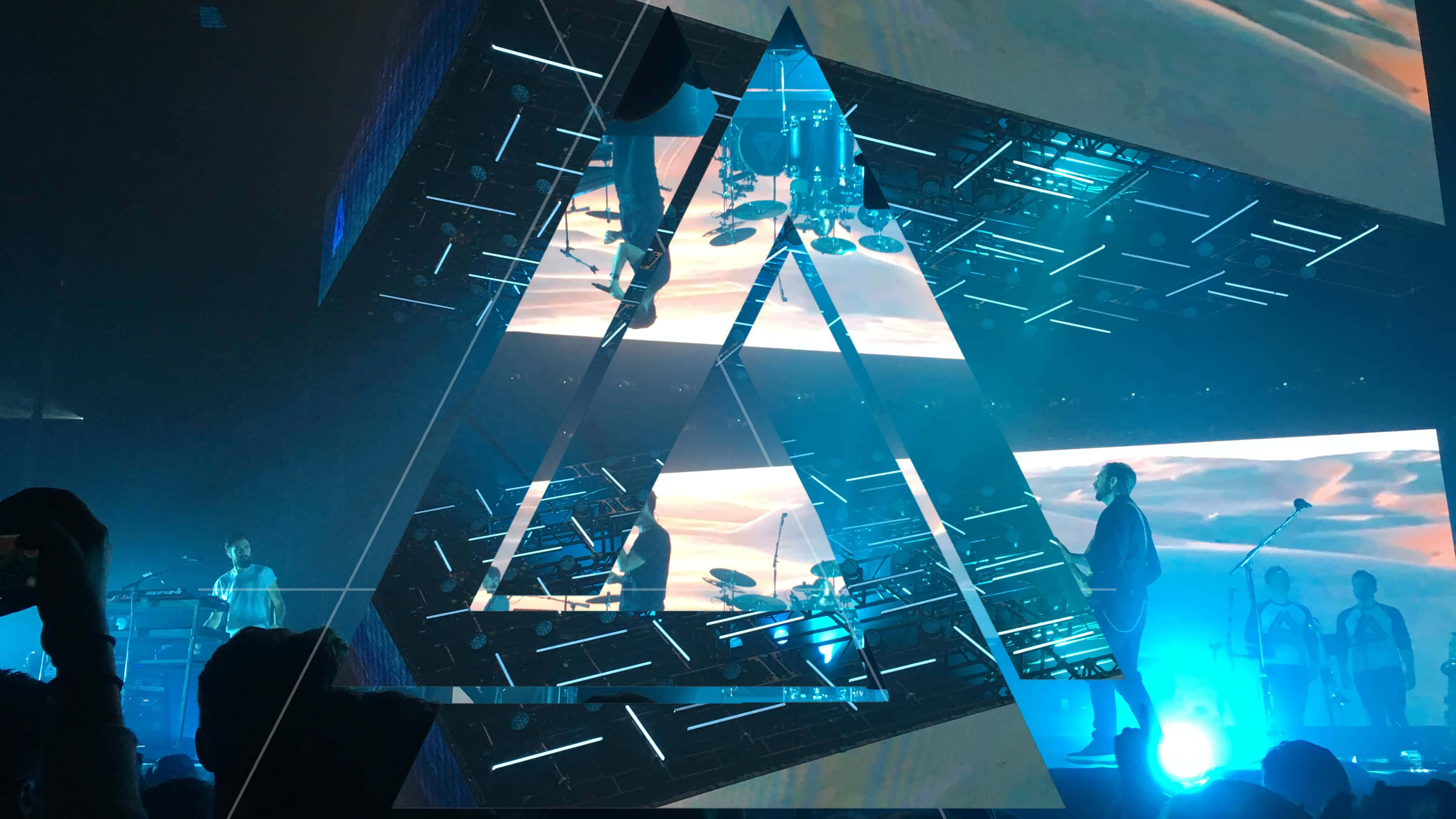 Bastille 桌布 壁紙 — 一個沐浴在藍光中的現場音樂會舞台，音樂家在幾何三角形燈光結構下表演，反射出日落天空。