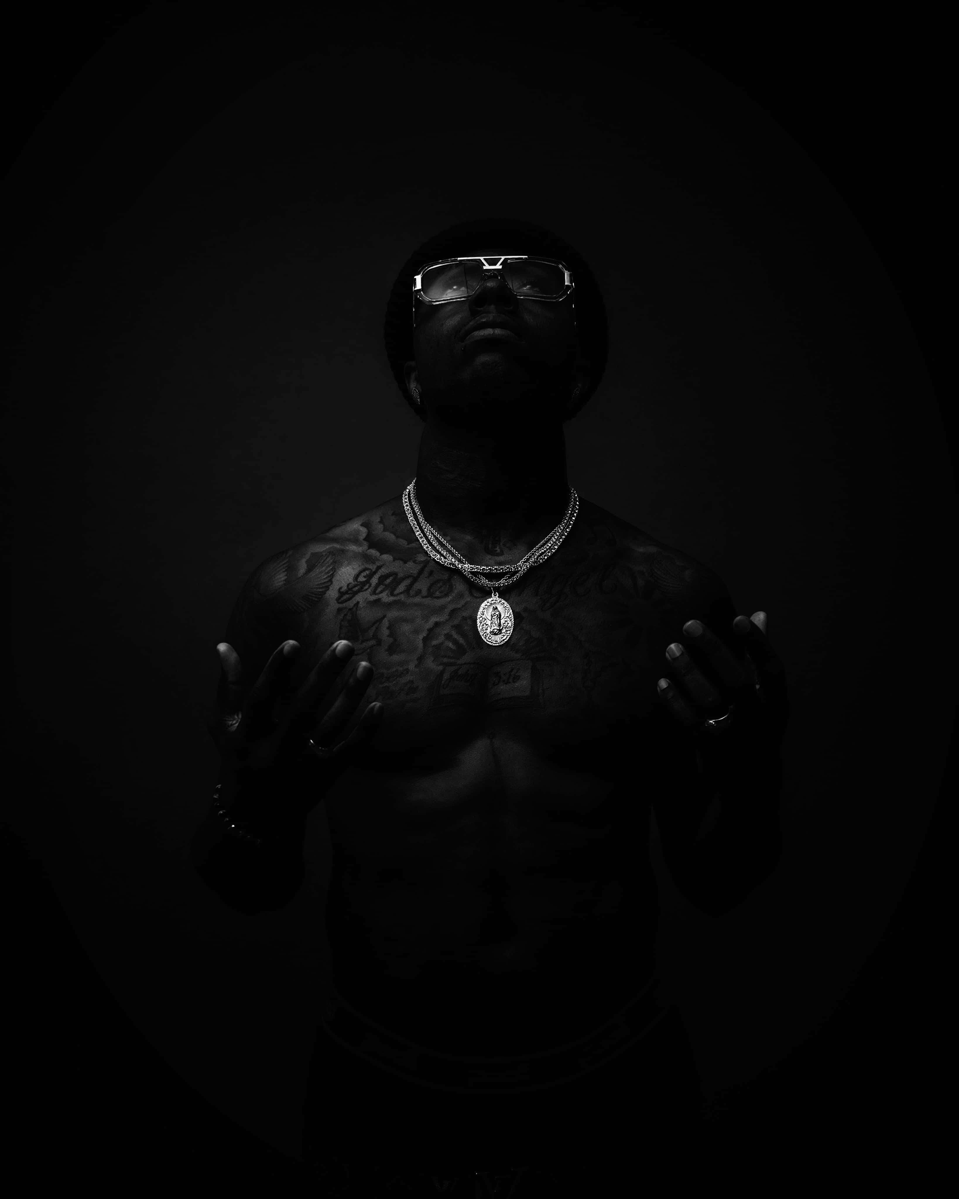 Rap Aesthetic 桌布 壁紙 — 一個沒穿上衣、有刺青且戴眼鏡的男人站在黑暗中，舉起雙手，形成一個高對比度的黑白戲劇性肖像。