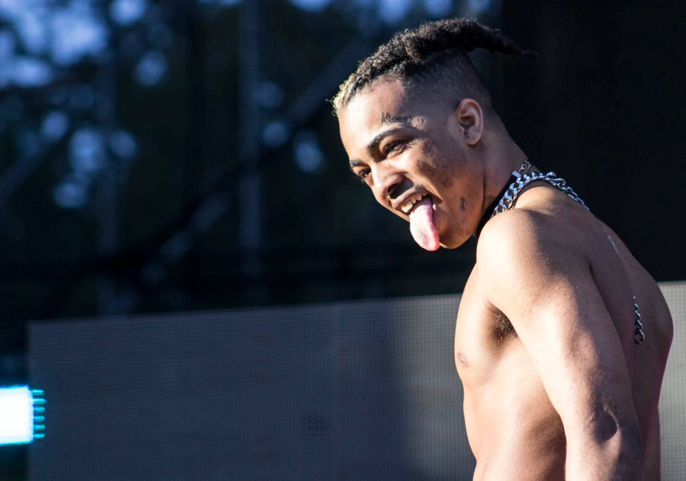 xxxtentacion 電腦 桌布 壁紙 — 一個沒穿上衣、綁辮子的年輕男子在明亮的舞台燈光下頑皮地伸出舌頭。