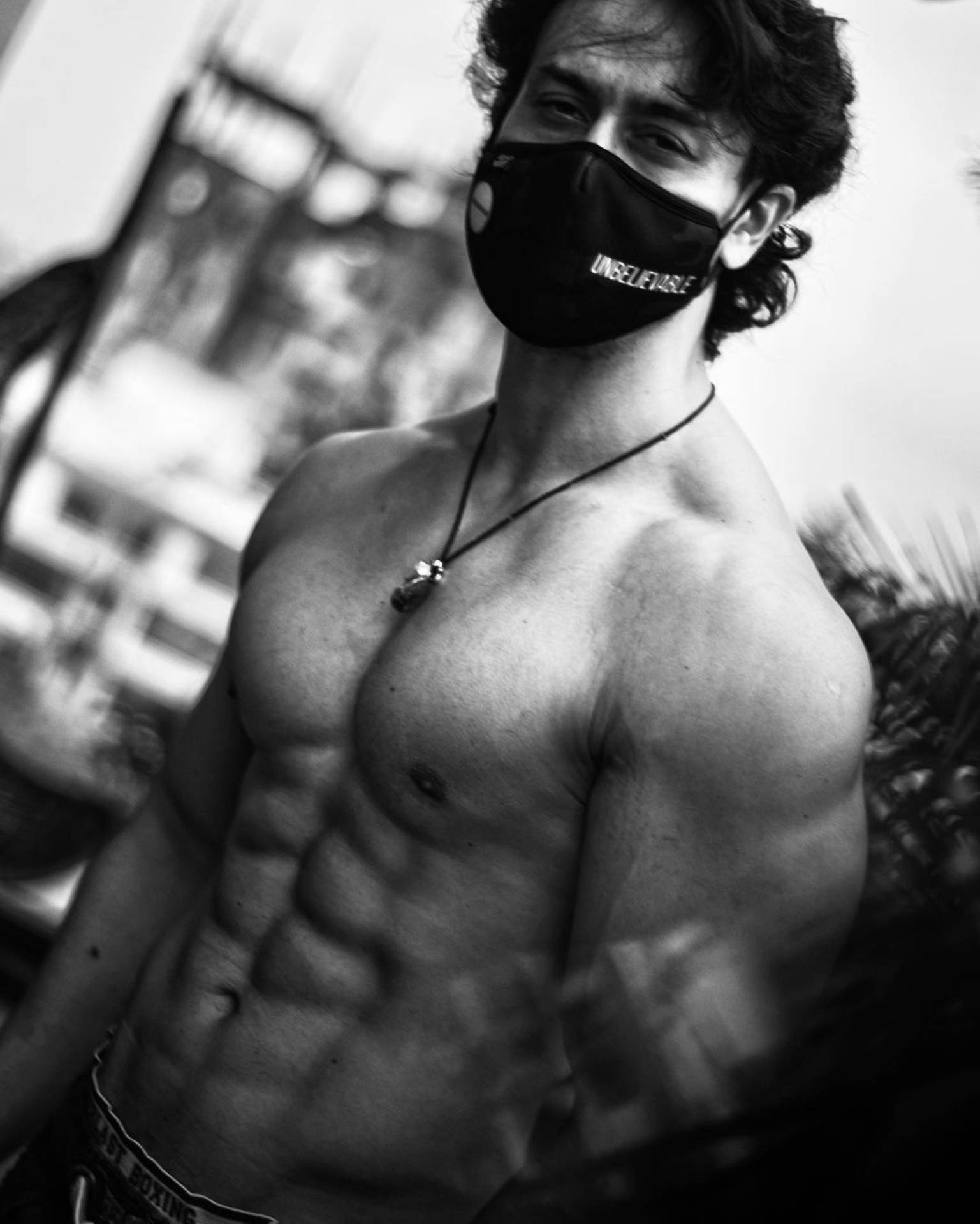 Tiger Shroff 桌布 壁紙 — 一個沒穿上衣、肌肉發達、捲髮的男人，戴著黑色口罩和項鍊，在戶外以黑白風格擺姿勢。