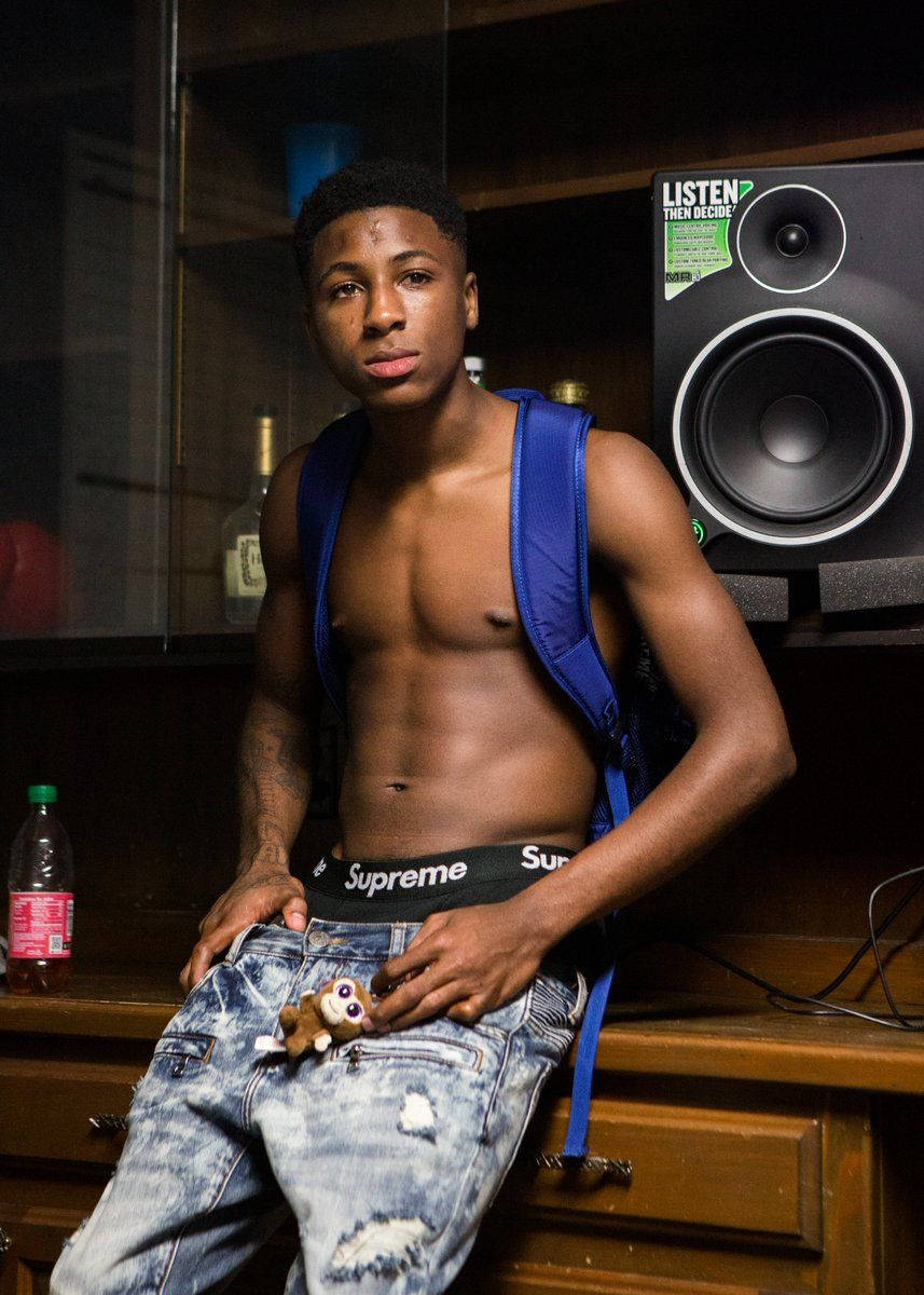 Youngboy 桌布 壁紙 — 一個沒穿上衣的年輕男子，背著藍色背包，靠在一張木桌旁，旁邊有一個大喇叭和一個瓶子。