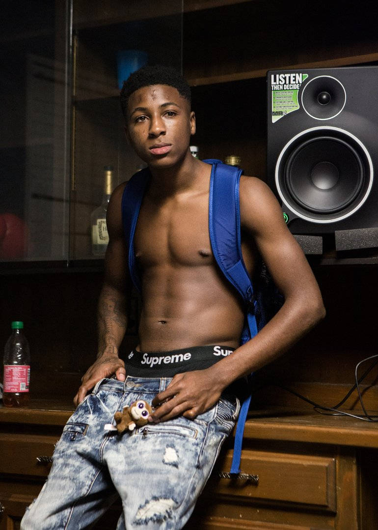 Youngboy 桌布 壁紙 — 一個沒穿上衣的年輕男子背著藍色背包，在昏暗的房間裡靠著一個木櫃，旁邊有一個大喇叭。