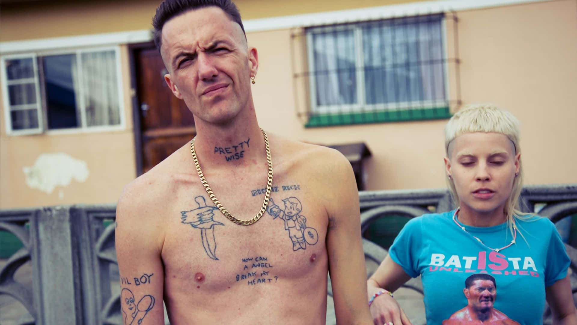 Tattoo 桌布 壁紙 — 一個沒穿上衣的男人，有刺青和金鍊，做了個苦臉，而一個金髮女人穿著藍色Batista T恤站在他旁邊。