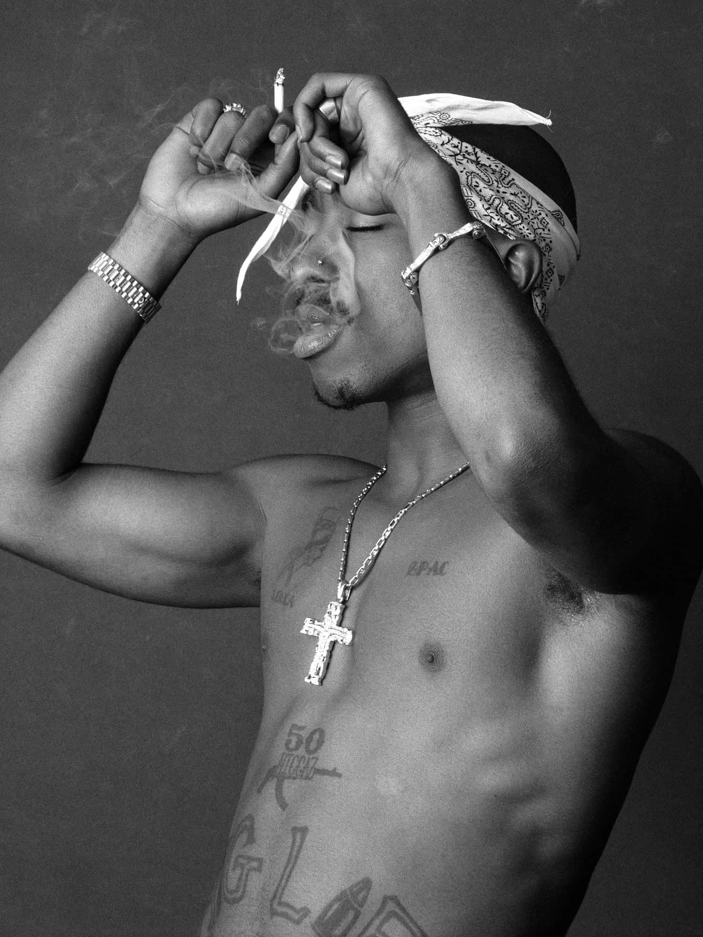tupac 桌布 壁紙 — 一個沒穿上衣的男人，有刺青和頭巾，在灰色背景前抽菸，黑白照片。