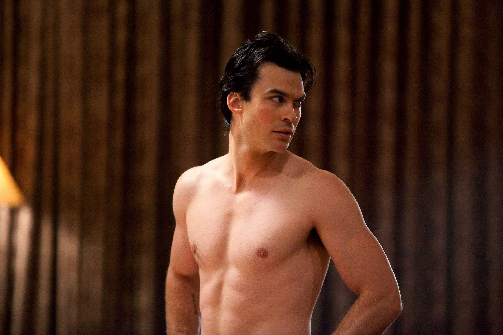Damon Salvatore 桌布 壁紙 — 一個沒穿上衣的肌肉男，深色頭髮，站在一個溫暖光線的房間，後面有木製窗簾。