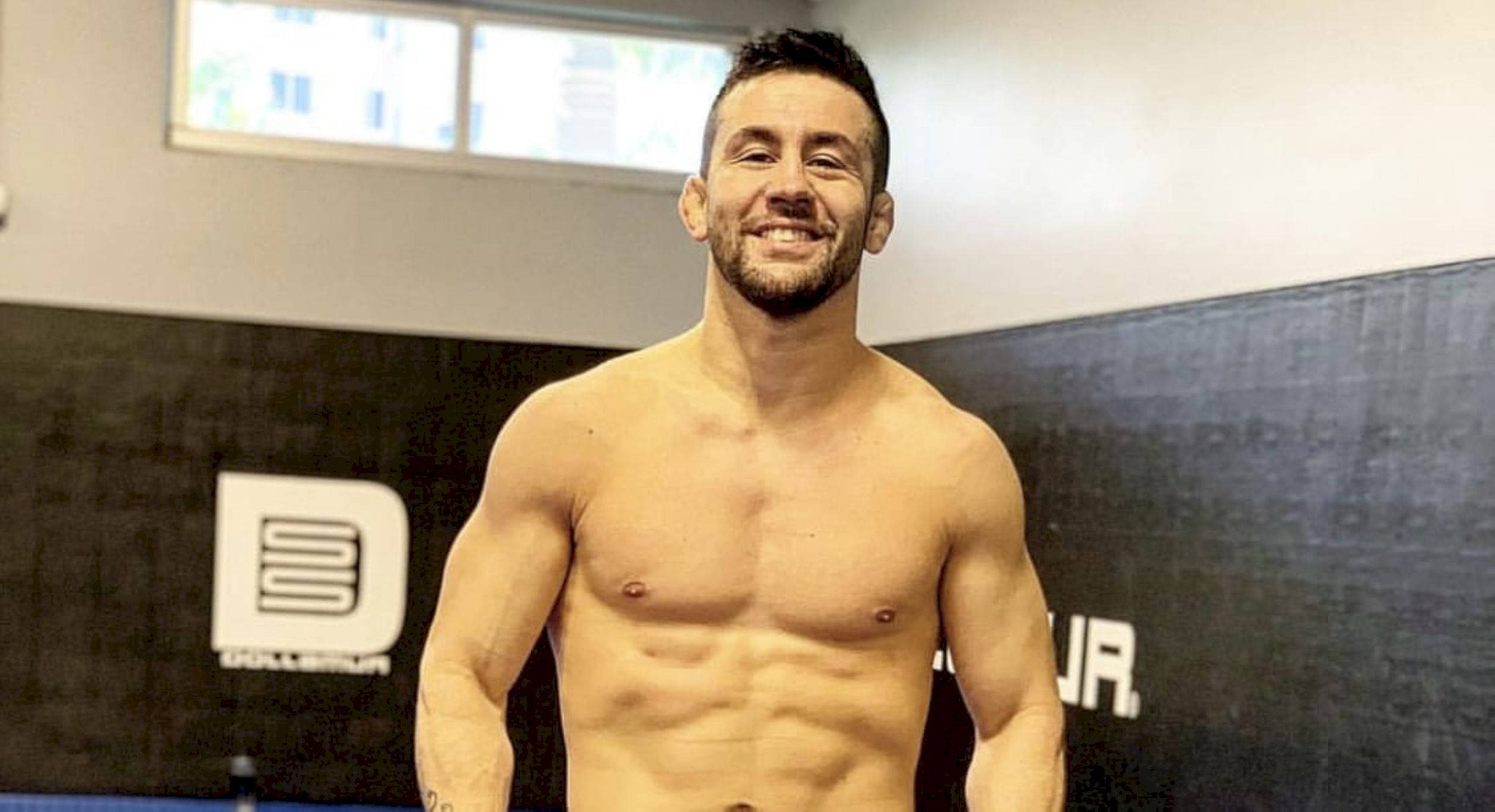 MMA 桌布 壁紙 — 一個沒穿上衣的鬍鬚肌肉男在黑色軟墊牆和標誌的健身房中自信地微笑。