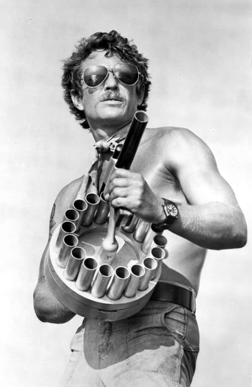 Tom Berenger 桌布 壁紙 — 一個沒穿衣服的男人戴著太陽眼鏡和鬍子，在普通天空背景下拿著一把大型多管武器。