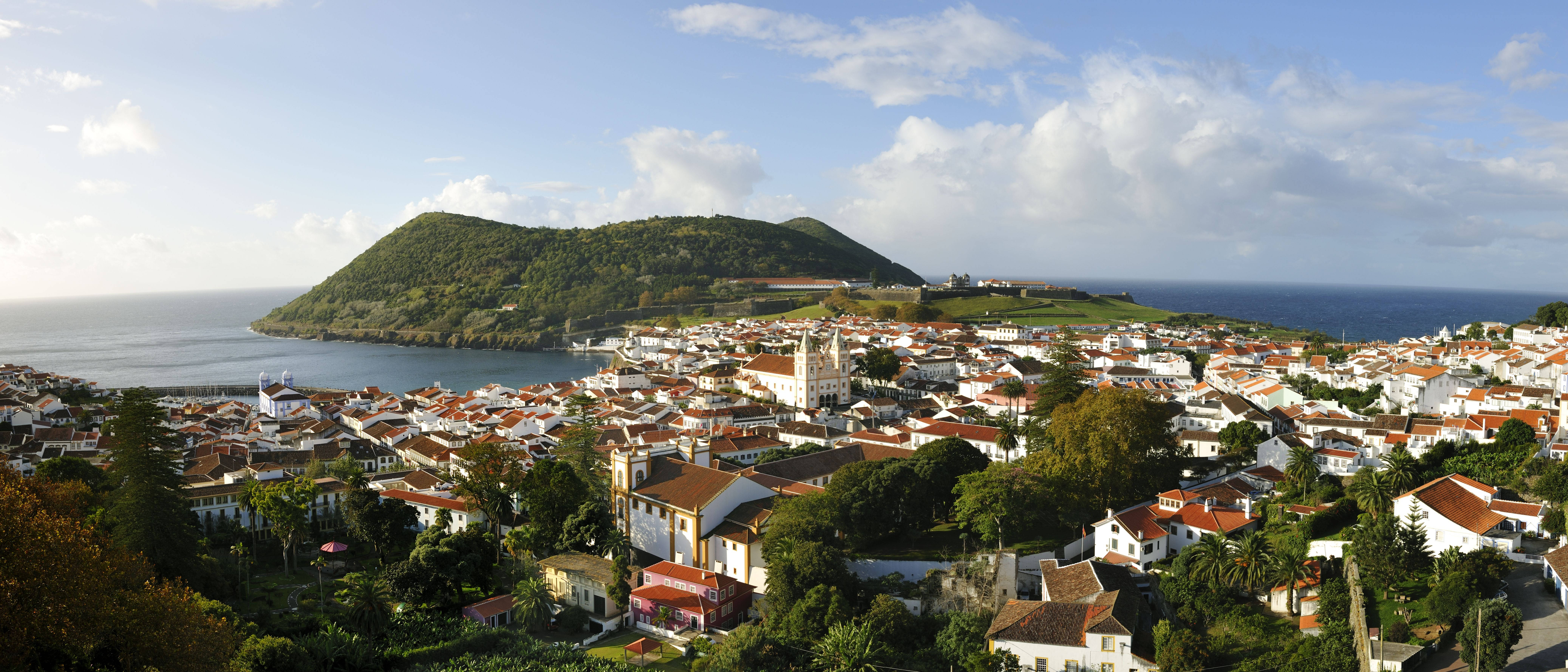 Portugal 桌布 壁紙 — 一個沿海小鎮，白色建築和紅色屋頂依附在綠色的山坡上，俯瞰著平靜的藍色大海，天空部分多雲。