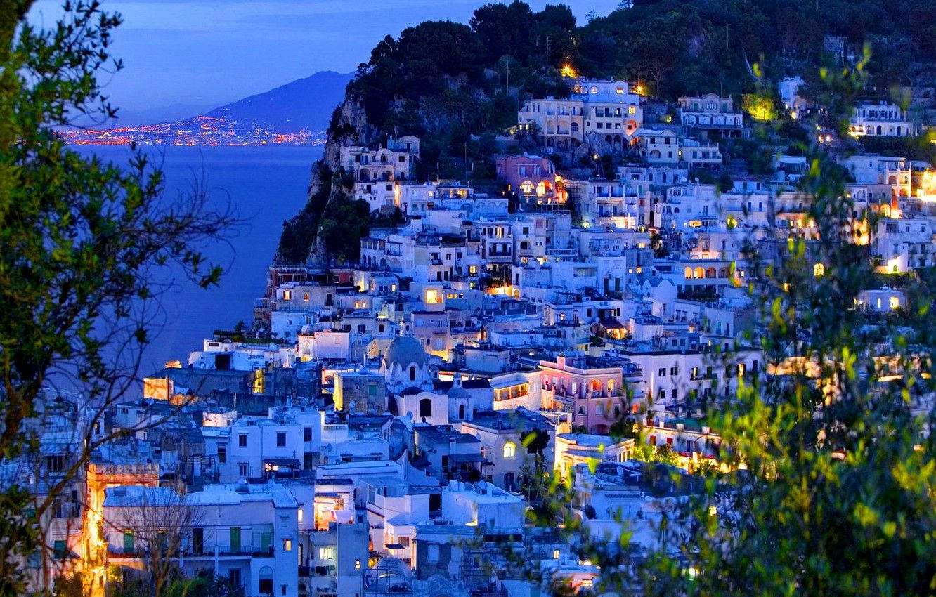 Capri 桌布 壁紙 — 一個沿海小鎮在黃昏時依附在懸崖邊，窗戶閃爍著光芒，下方是平靜的海面。