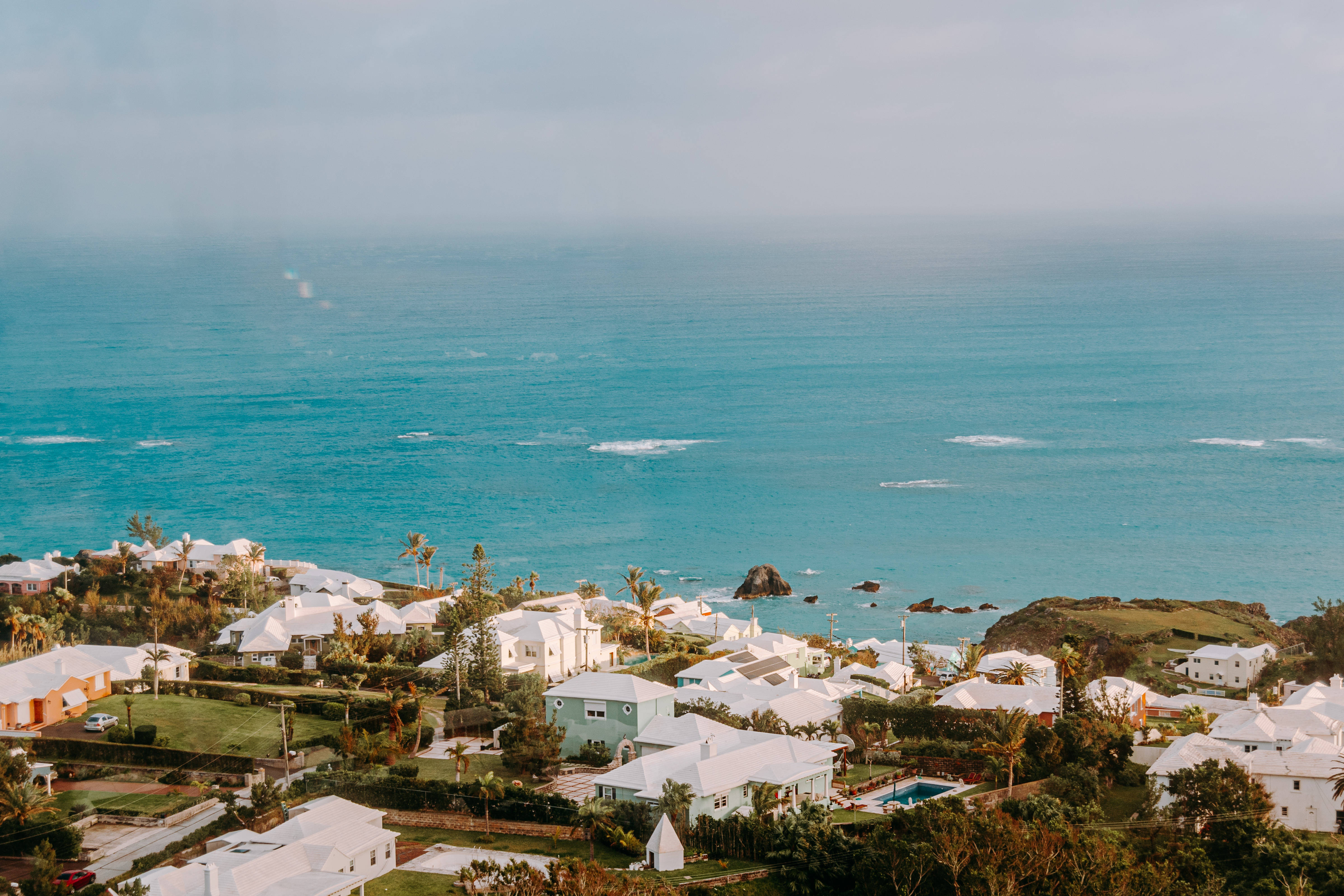 Bermuda 桌布 壁紙 — 一個沿海村莊，白色房屋隱藏在綠樹叢中，俯瞰著廣闊的碧綠海洋，天空朦朧。