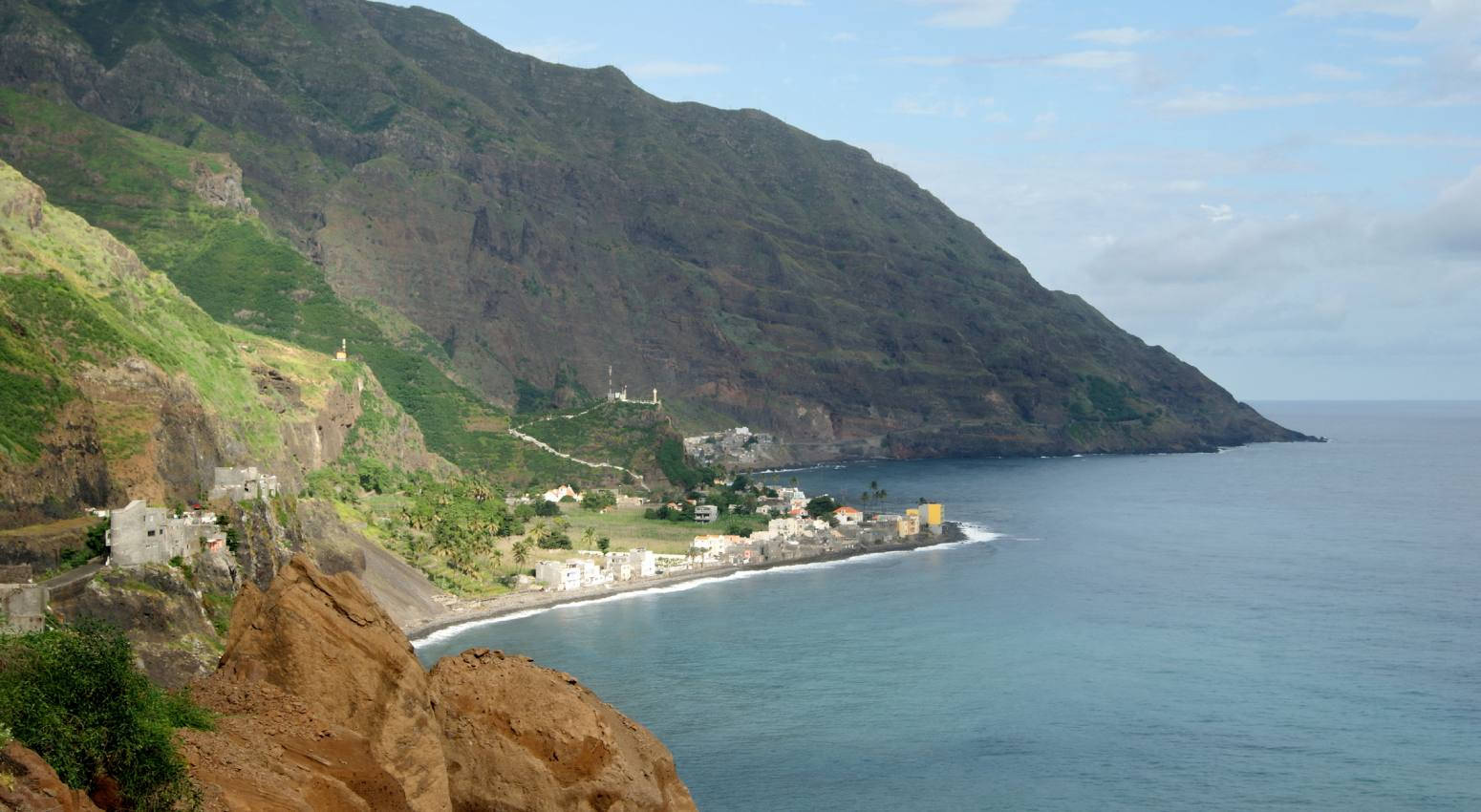 島嶼 桌布 壁紙 — 一個沿海村莊依附在陡峭的綠色山邊，旁邊是碧綠的海水，天空部分多雲。