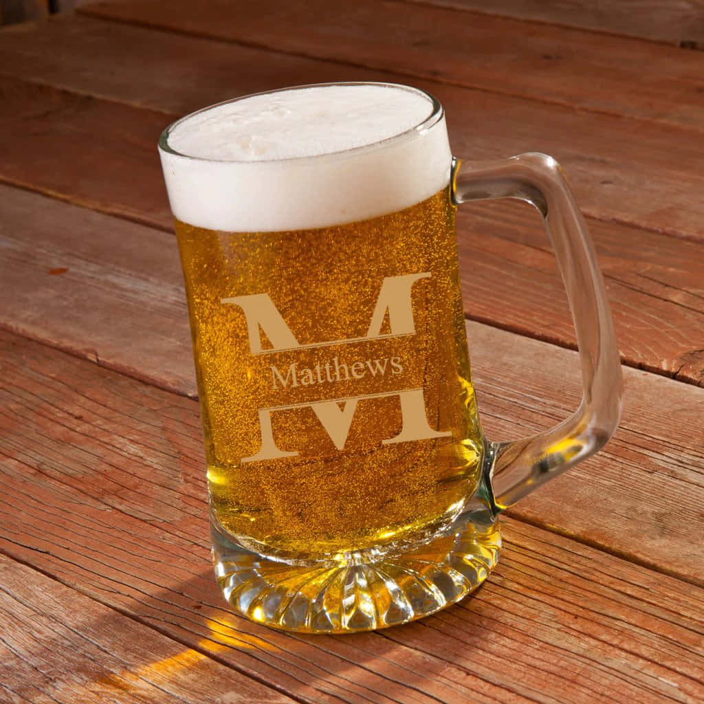 Beer 桌布 壁紙 — 一個泡沫豐富的金色啤酒杯，上面刻有「Matthews」，放在一張飽經風霜的木桌上，沐浴在溫暖的陽光中。