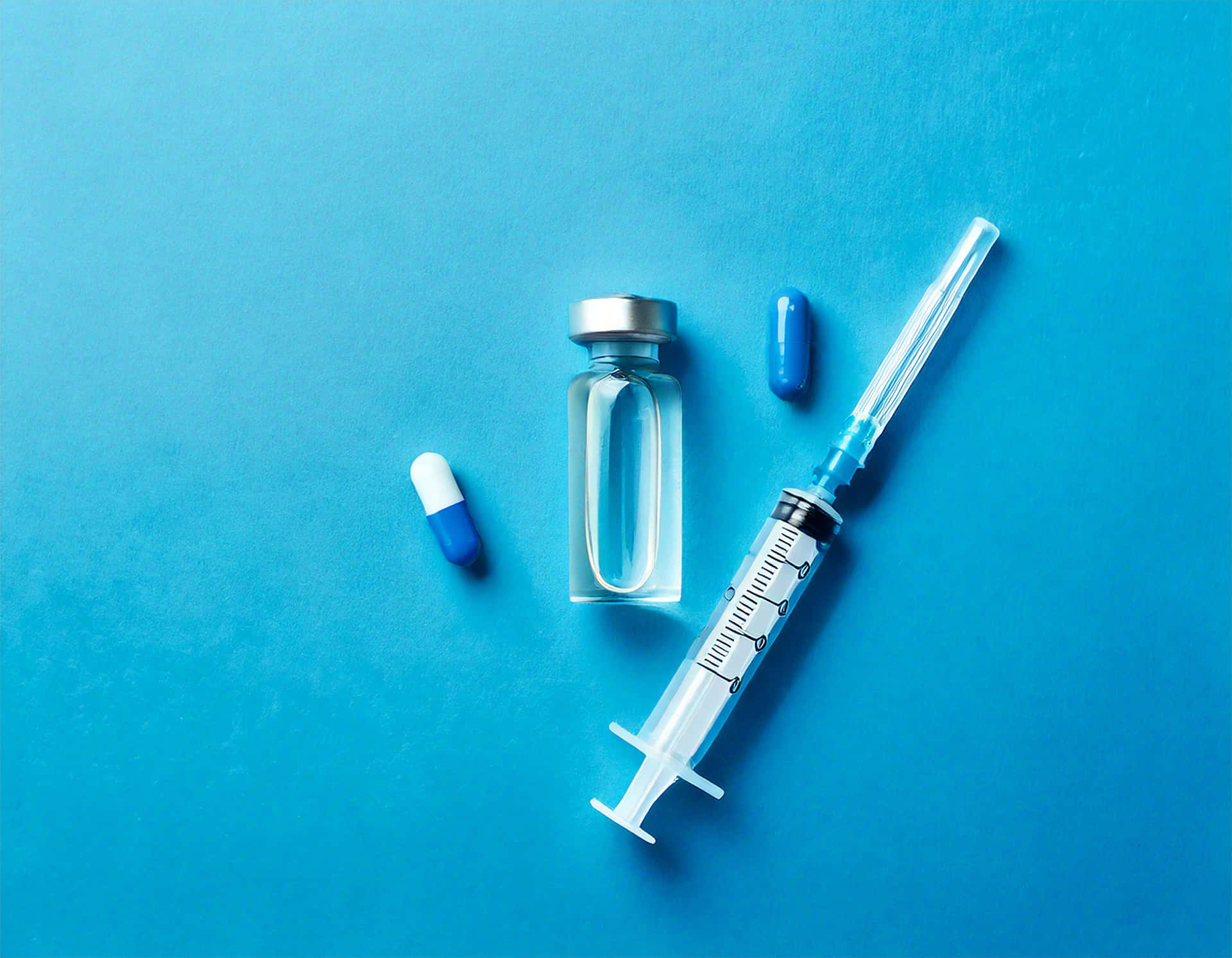 Syringe and Capsule 壁紙 — 一個注射器藥瓶和兩粒膠囊放在純藍色表面上，構成簡潔極簡的構圖。