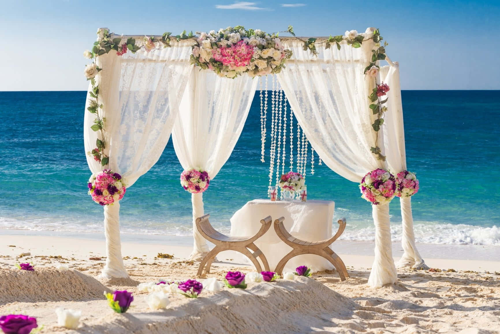 Wedding 桌布 壁紙 — 一個浪漫的海灘婚禮拱門，裝飾著粉紅色和白色的花朵以及薄紗窗簾，矗立在金色沙灘上，旁邊是碧綠的海浪，頭頂是清澈的藍天。