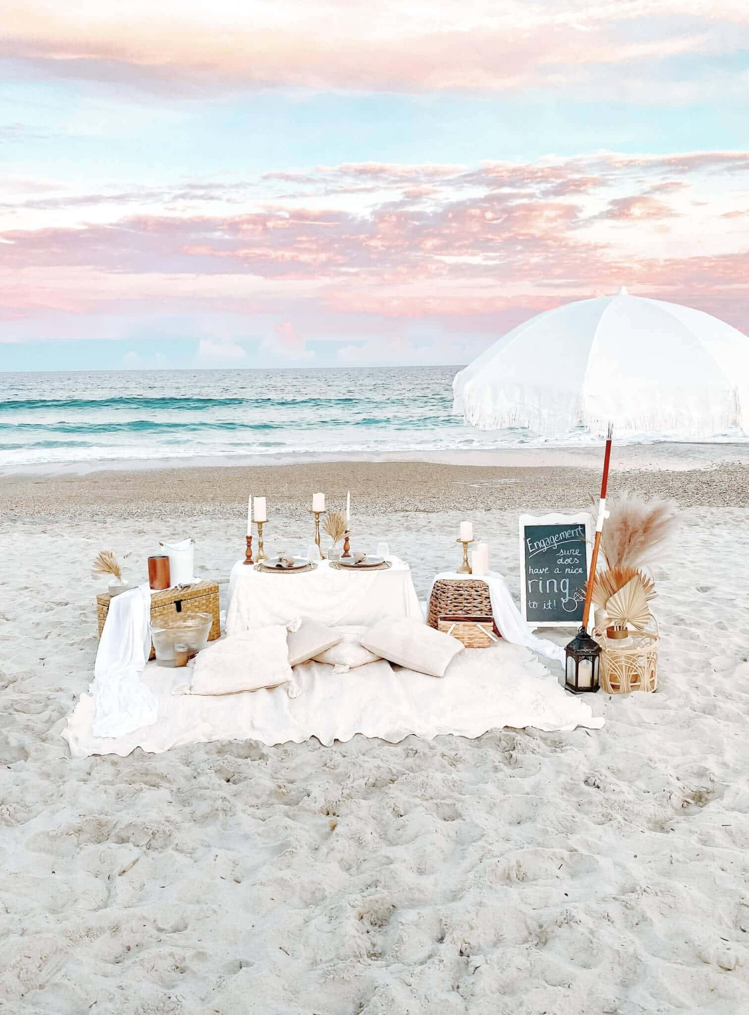 Beach Picnic 桌布 壁紙 — 一個浪漫的海灘野餐佈置，配有白色桌布和遮陽傘，在柔和的粉紅色和藍色日落天空下。