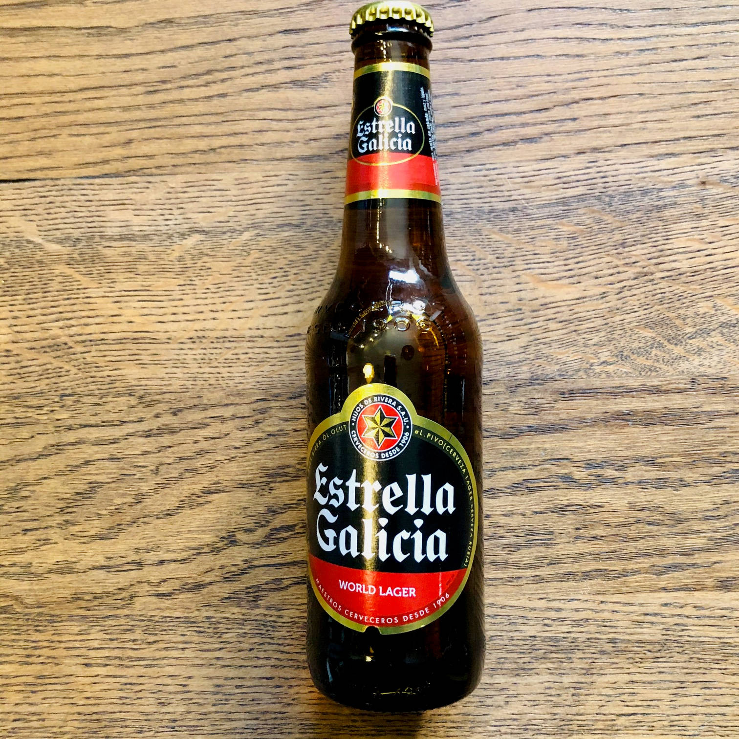 alcohol 桌布 壁紙 — 一個深棕色的Estrella Galicia啤酒瓶，帶有紅色和金色標籤，直立放在有紋理的木質表面上。