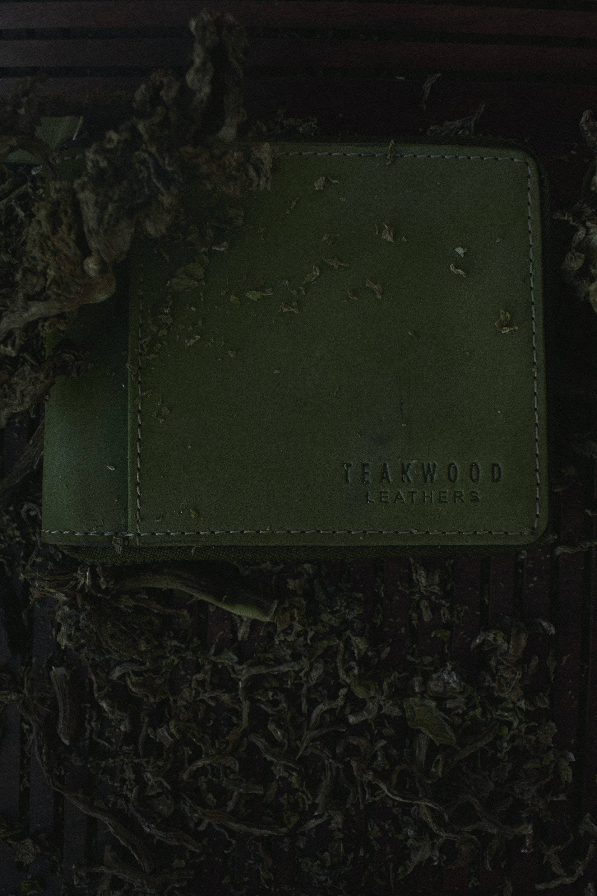 black aesthetic tumblr iPhone 桌布 壁紙 — 一個深橄欖綠色皮革錢包，印有 'TEAKWOOD LEATHERS' 字樣，靜靜地躺在陰影中的乾葉堆上。