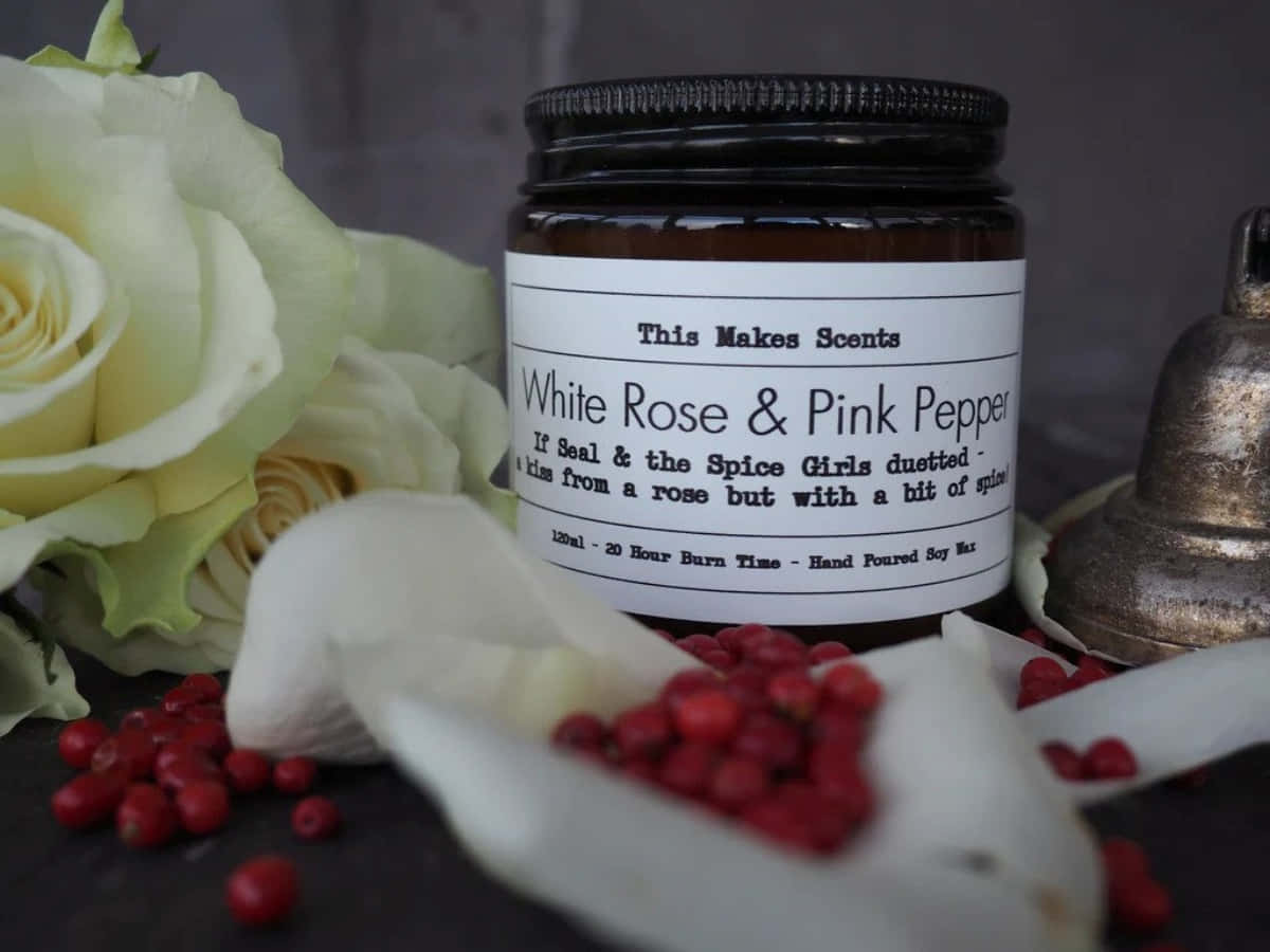 Pink Peppercorn 桌布 壁紙 — 一個深琥珀色蠟燭罐，標籤為 White Rose & Pink Pepper，旁邊有淡黃色玫瑰和散落的紅胡椒粒，放在深色表面上。