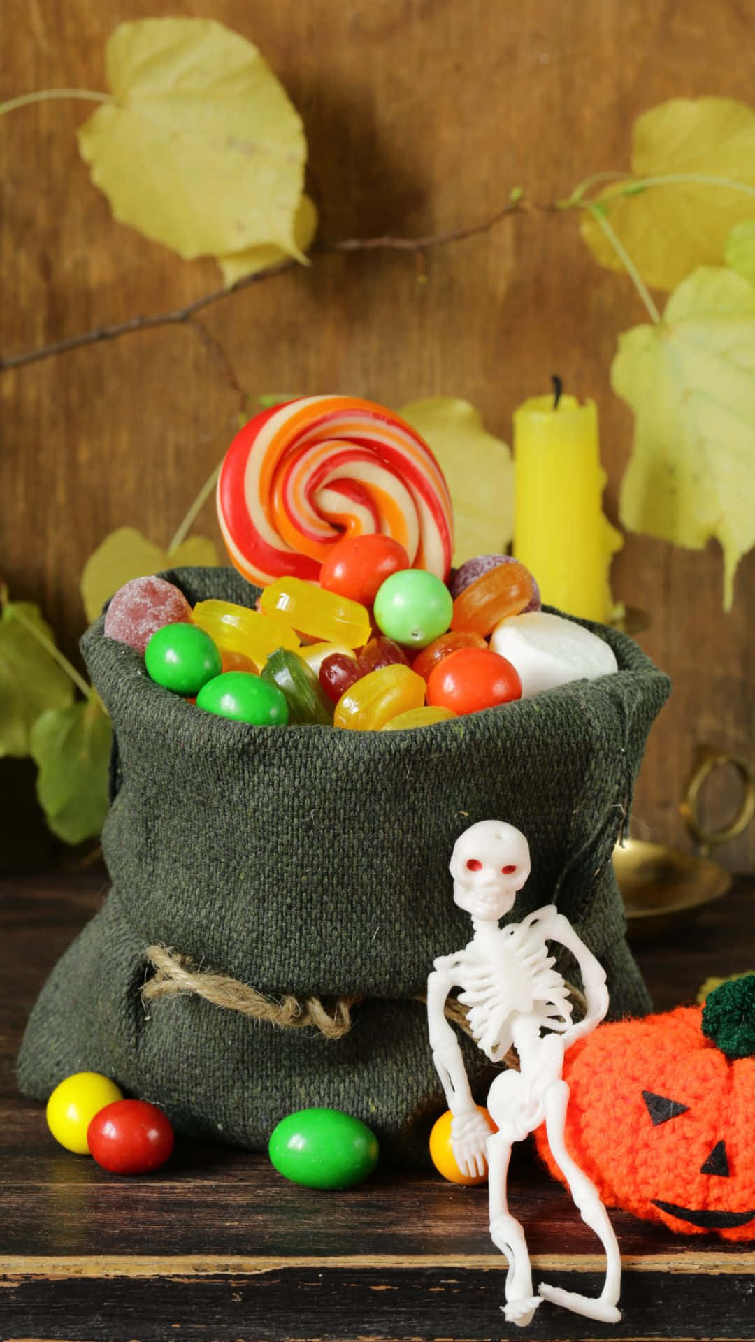 Trick Or Treat Bags 桌布 壁紙 — 一個深綠色袋子，裝滿了色彩繽紛的糖果和一個巨大的螺旋棒棒糖，旁邊放著一個小骷髏和一個編織南瓜，背景是質樸的木頭和秋天的樹葉。