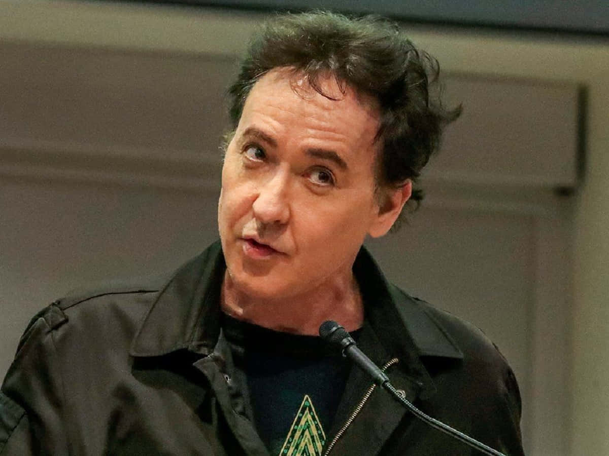 John Cusack 桌布 壁紙 — 一個深色亂髮的男人對著麥克風說話，身穿黑色外套，內搭帶有綠色幾何圖案的深色襯衫。