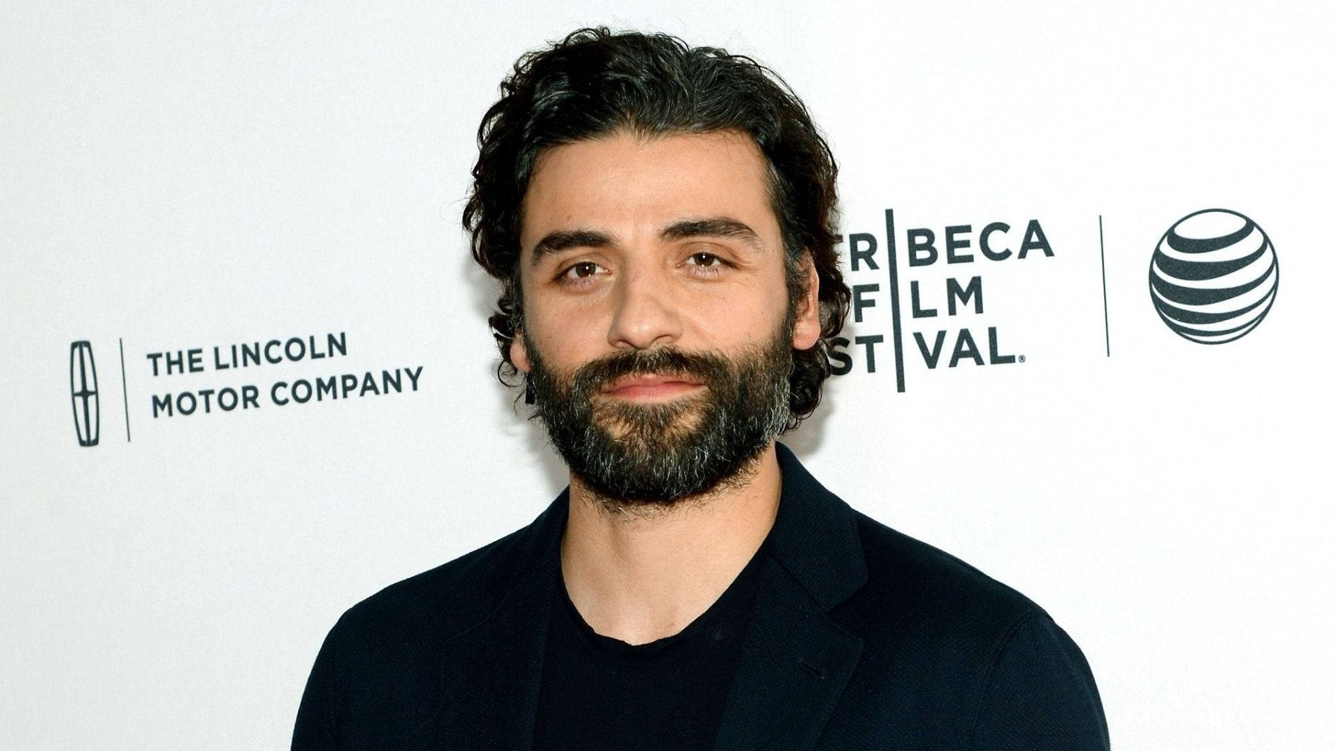 oscar 桌布 壁紙 — 一個深色捲髮、留著大鬍子的男人在帶有標誌的白色背景前對著鏡頭微笑。