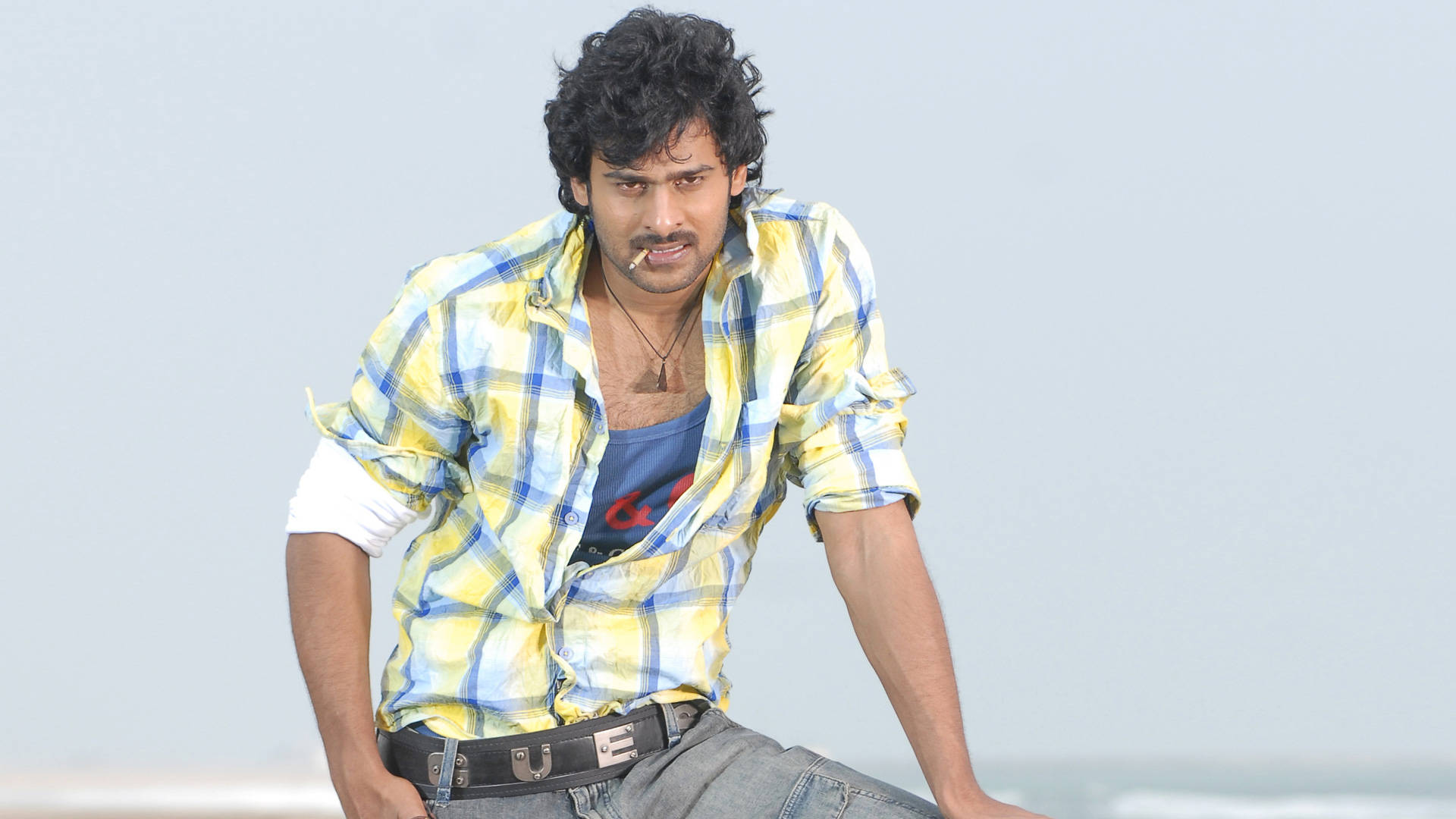 Prabhas HD 桌布 壁紙 — 一個深色捲髮、留鬍子的男人，穿著藍色背心外搭黃藍格紋襯衫，隨意地坐在沙灘上。