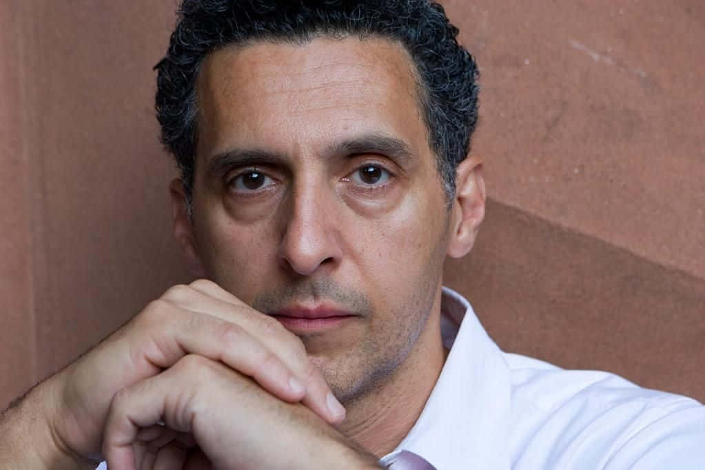 John Turturro 桌布 壁紙 — 一個深色捲髮、眼神銳利的男人，將下巴擱在交疊的雙手上，靠著質感豐富的赤陶牆。