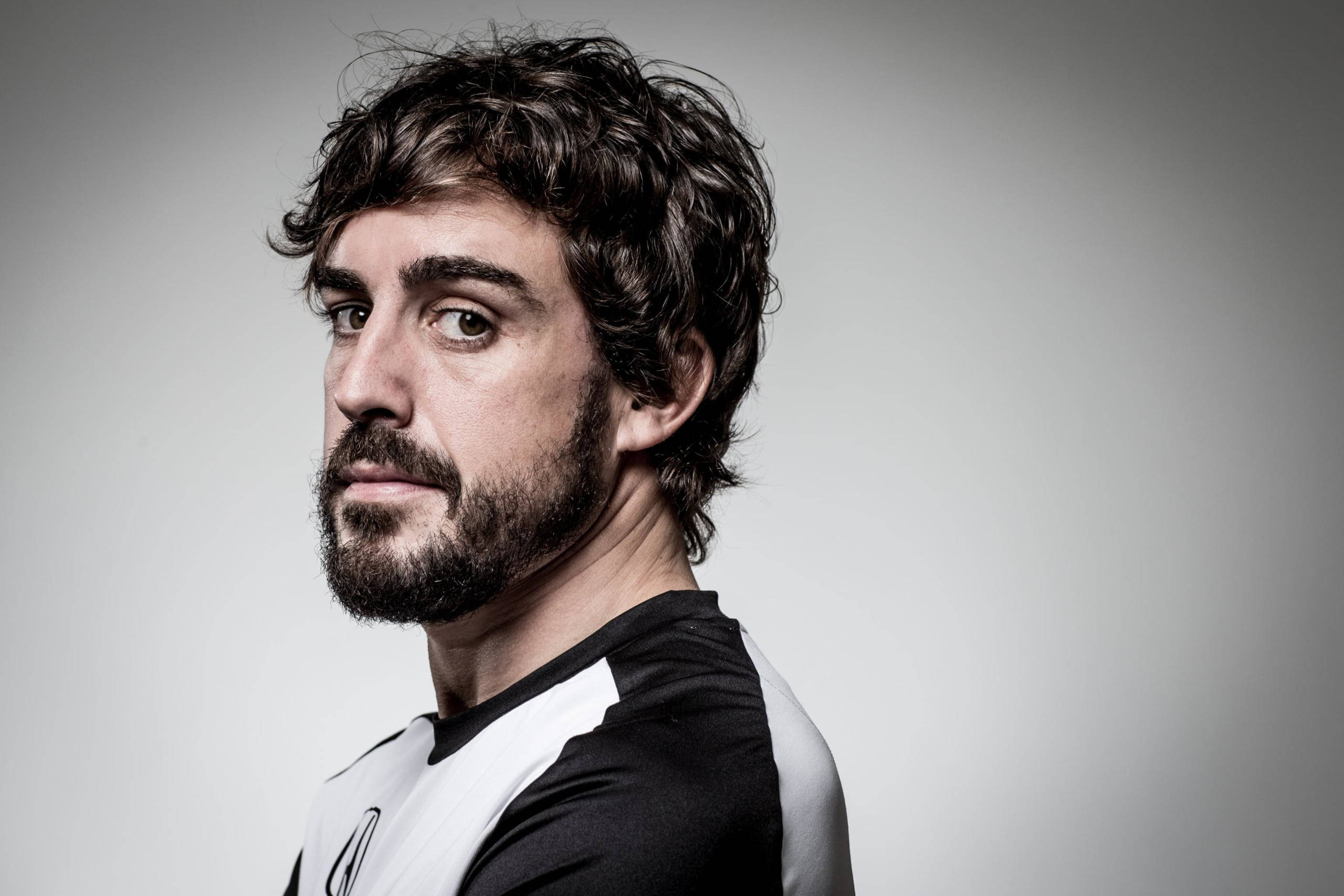 Fernando Alonso 桌布 壁紙 — 一個深色捲髮和鬍鬚的男人，在灰色中性工作室背景中，越過肩膀凝視著。