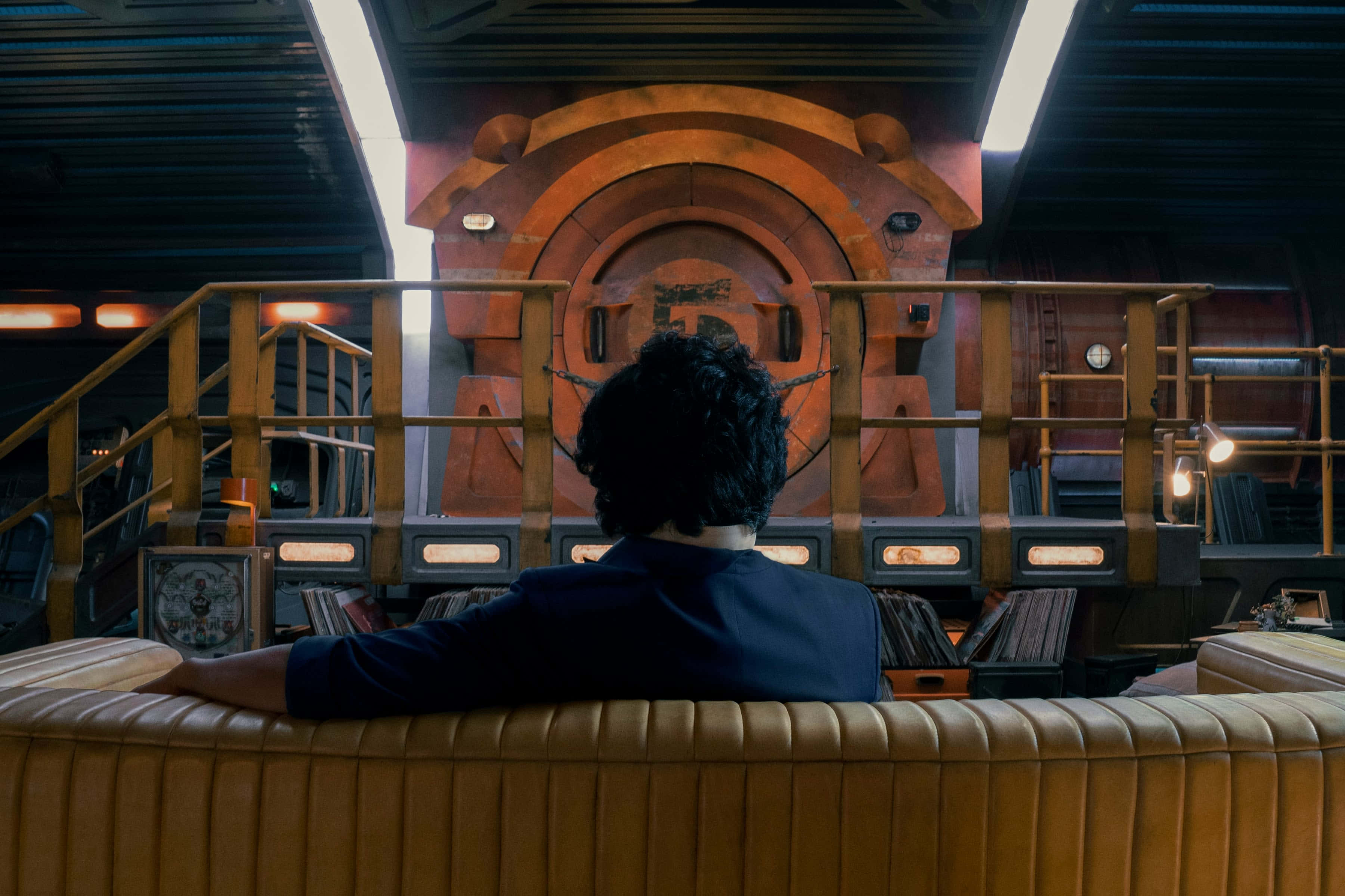 Spike Spiegel 桌布 壁紙 — 一個深色捲髮的人坐在棕色皮沙發上，面向一扇巨大的發光橙色圓形門，身處昏暗的科幻室內。