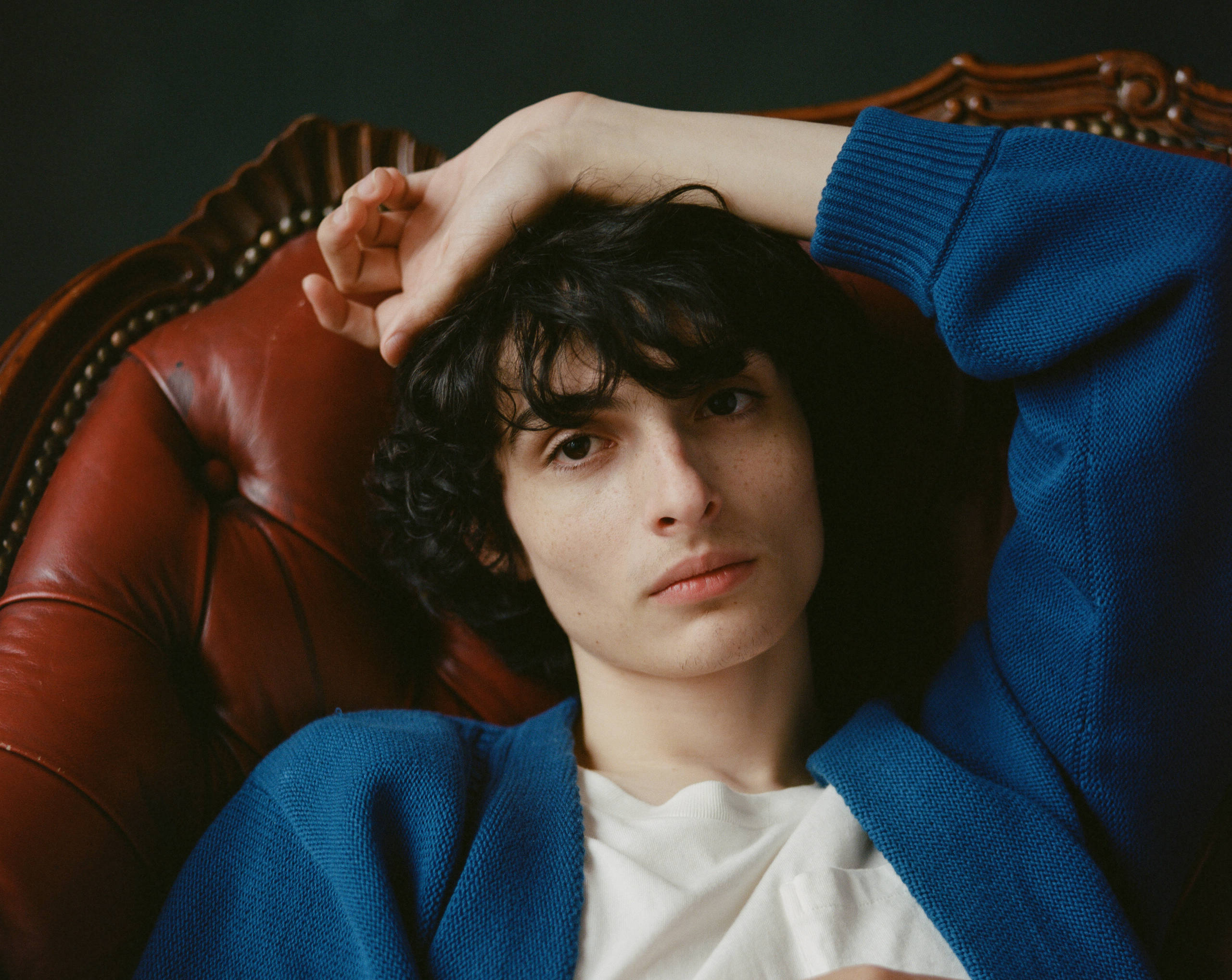 finn wolfhard 桌布 壁紙 — 一個深色捲髮的人穿著藍色毛衣，將頭靠在復古紅色皮椅上。