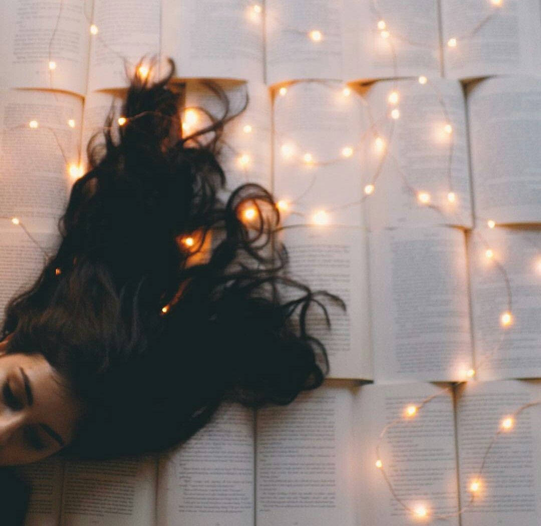 Fairy Lights Aesthetic 桌布 壁紙 — 一個深色捲髮的人靠在一面由溫暖串燈照亮的打開的書牆上休息。