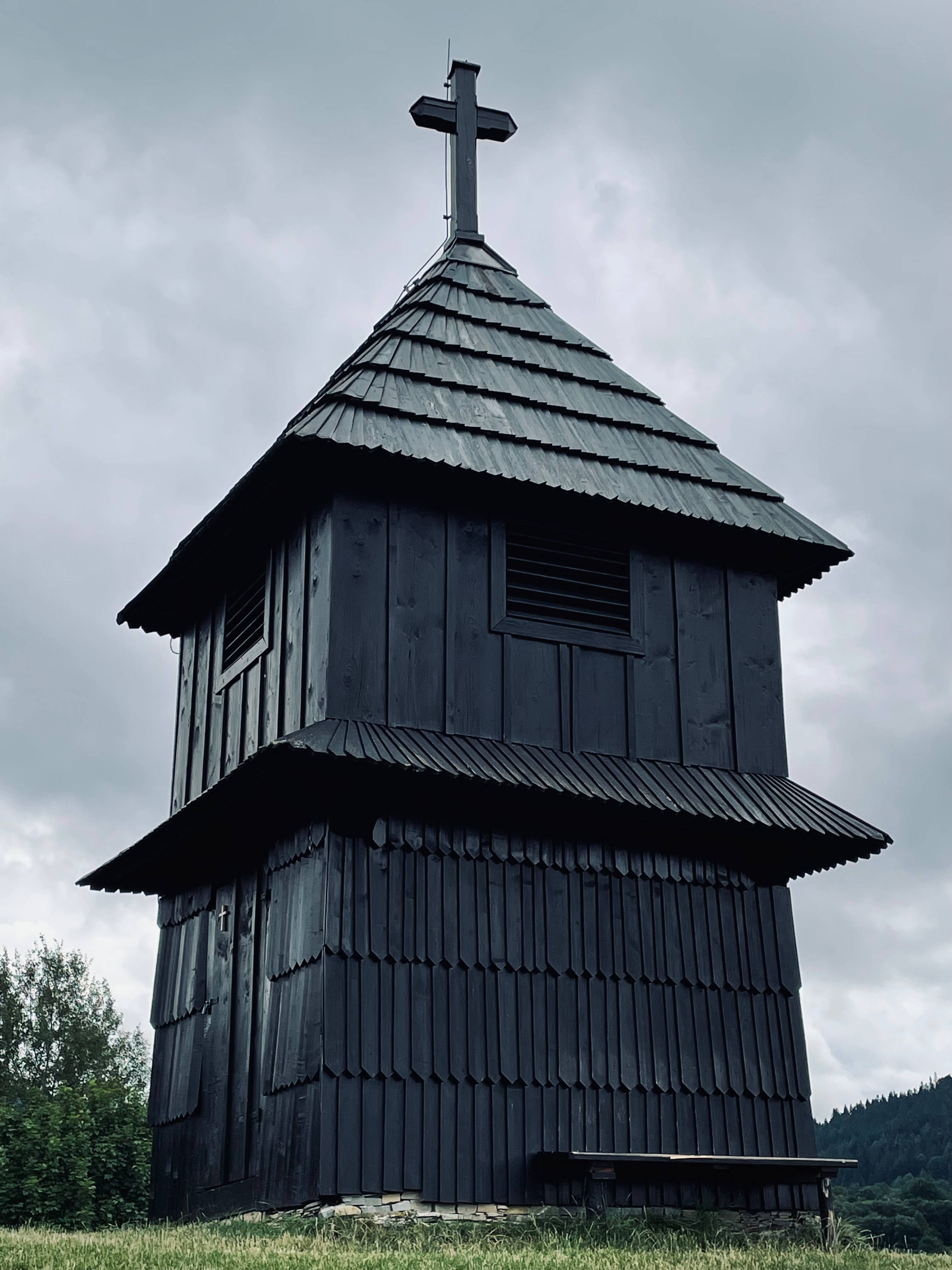 Slovakia 桌布 壁紙 — 一個深色木製教堂鐘樓帶有十字架，在陰天下的草地上莊嚴地矗立。
