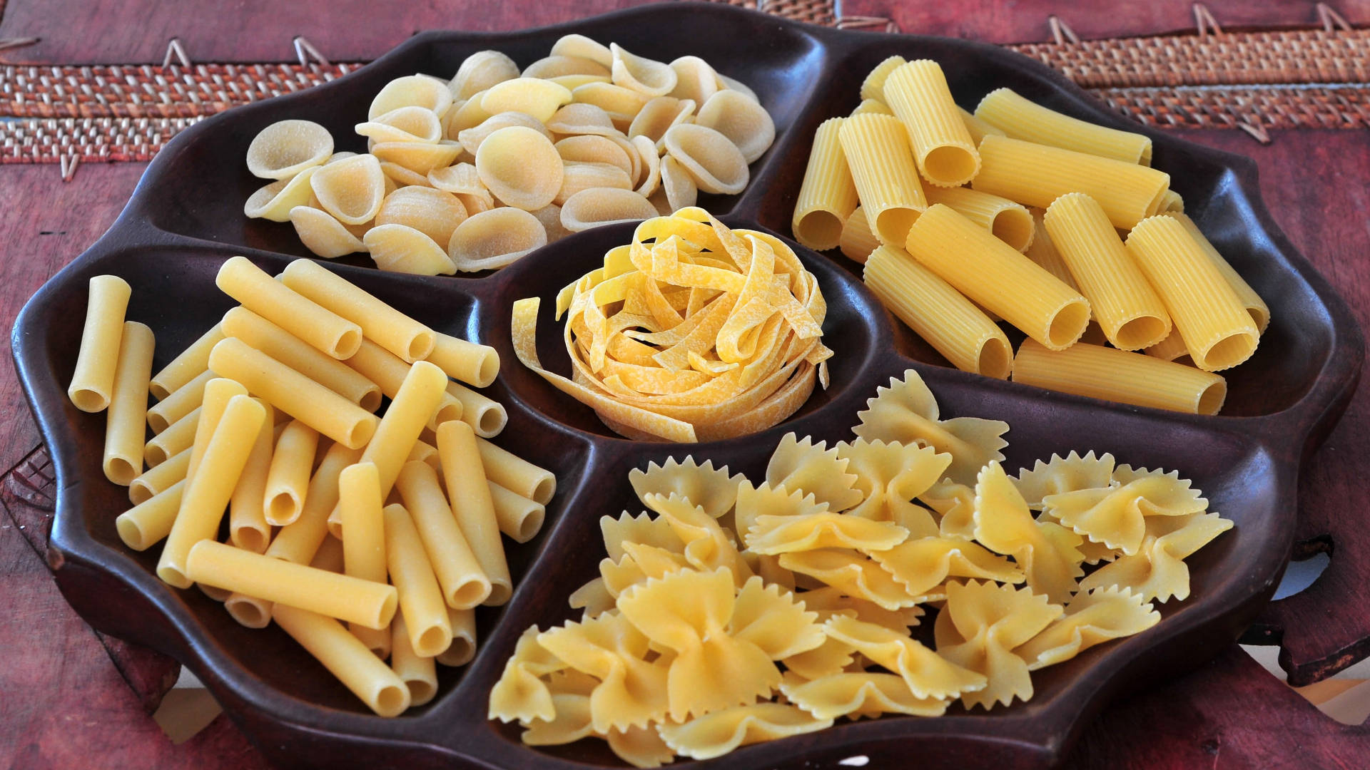 Pasta 桌布 壁紙 — 一個深色木製餐盤整齊地擺放著五種未煮熟的義大利麵。