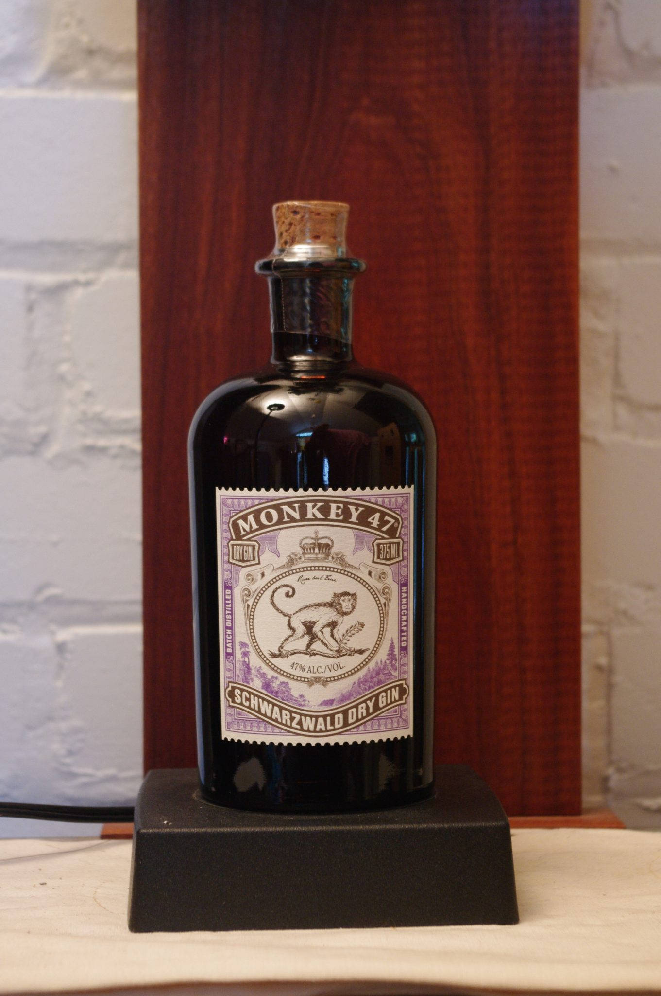 bottle 桌布 壁紙 — 一個深色玻璃瓶的Monkey 47 Schwarzwald Dry Gin，配有軟木塞，放在黑色支架上，背景是白色磚牆和木質面板。