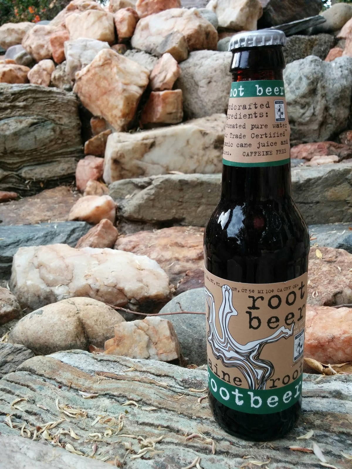 Drink 桌布 壁紙 — 一個深色玻璃瓶裝的root beer，帶有鄉村風格標籤，放在一條風化的木頭上，背景是堆疊的岩石。