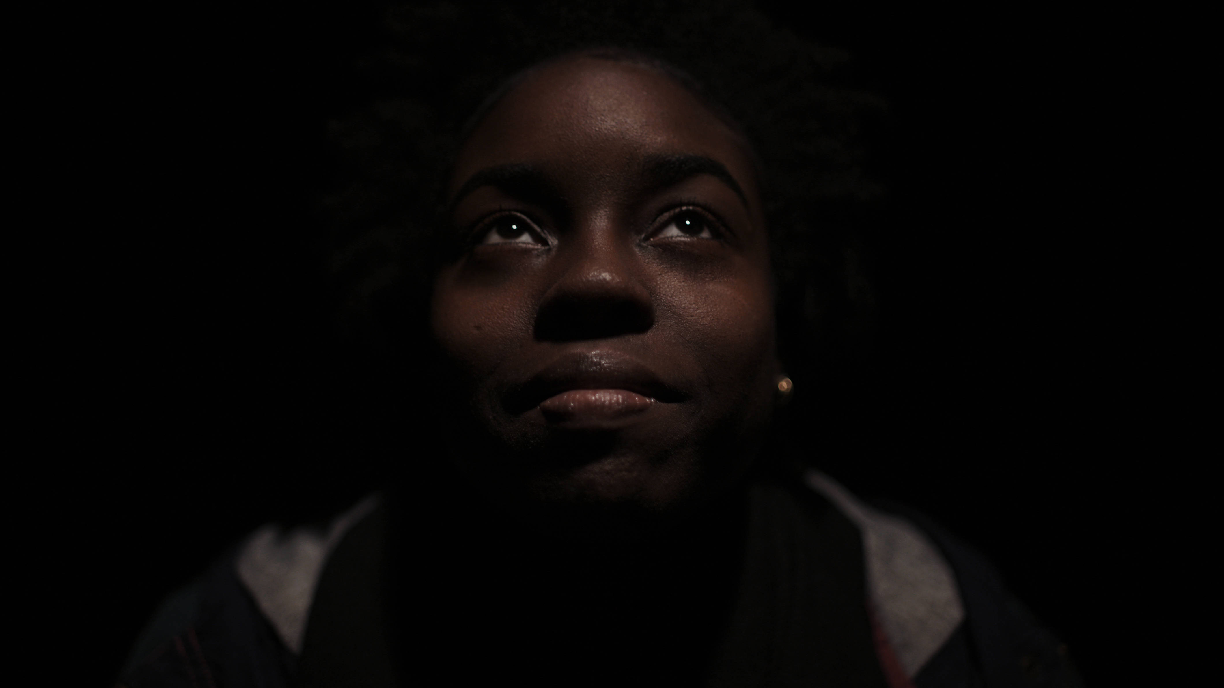 Black Girl 桌布 壁紙 — 一個深色皮膚、捲髮的人在昏暗環境中向上看，戲劇性的燈光突顯他們的臉部。