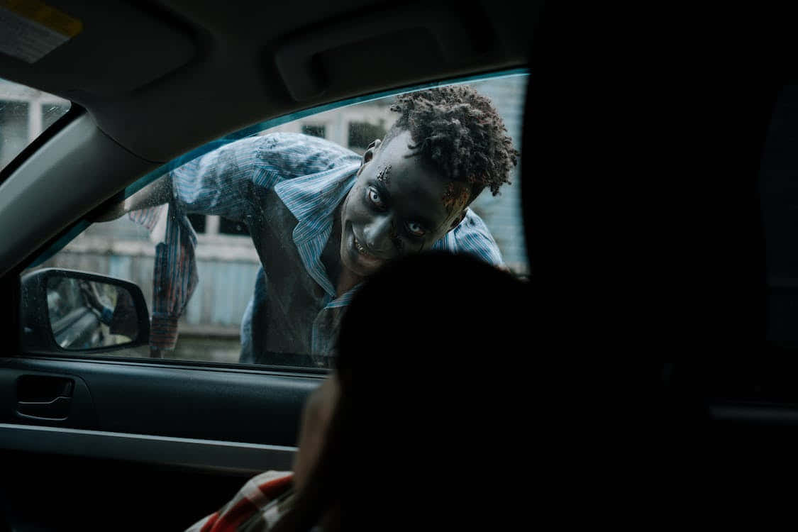 Scary Zombie 桌布 壁紙 — 一個深色皮膚、捲髮的人靠向車窗，露出燦爛的笑容，而另一個人則坐在陰影中的車內。