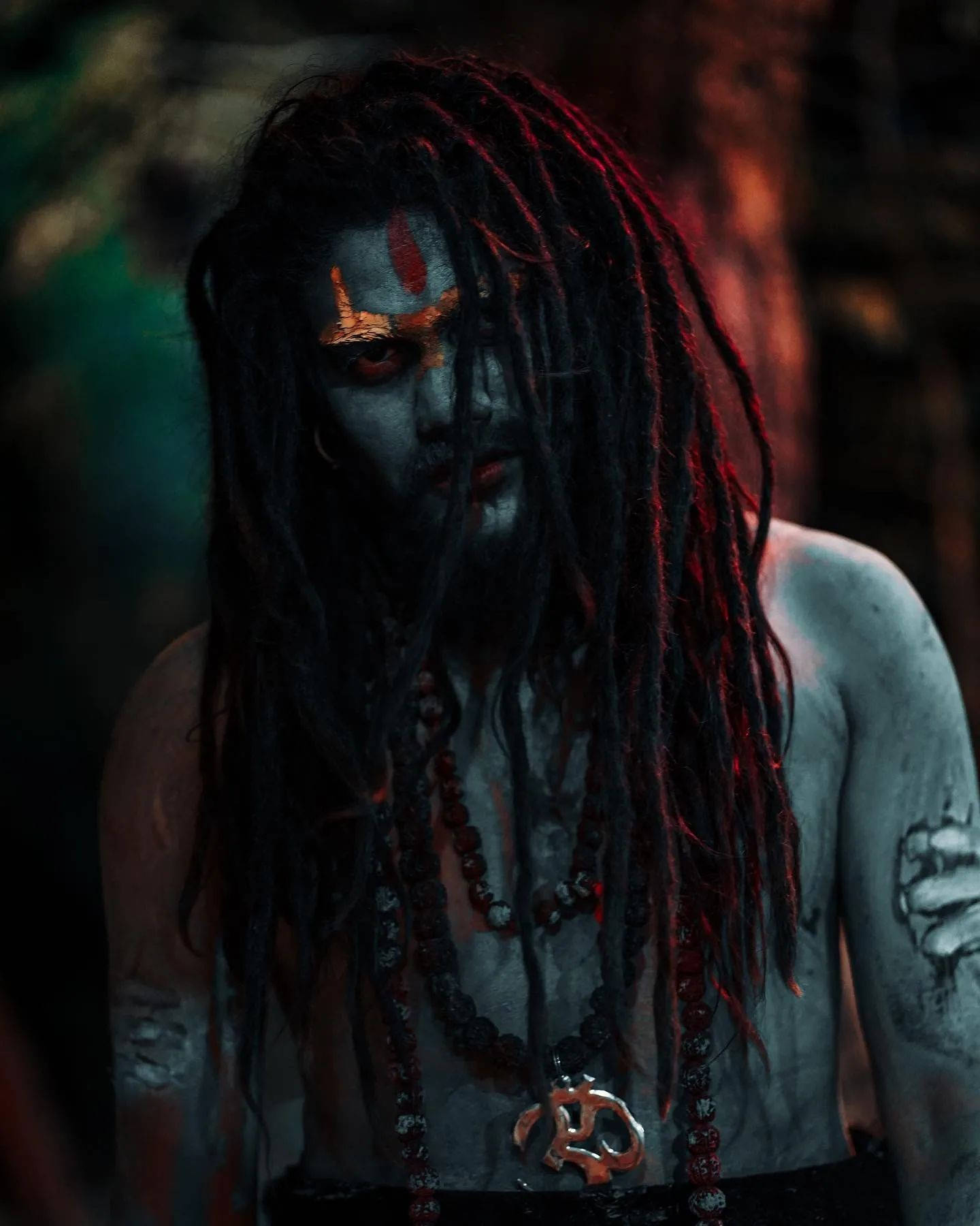 Aghori 桌布 壁紙 — 一個深色皮膚、梳著辮子、帶有紅色標記的人物，在陰暗的森林中，被詭異的紅色和綠色光芒照亮，凝視著。