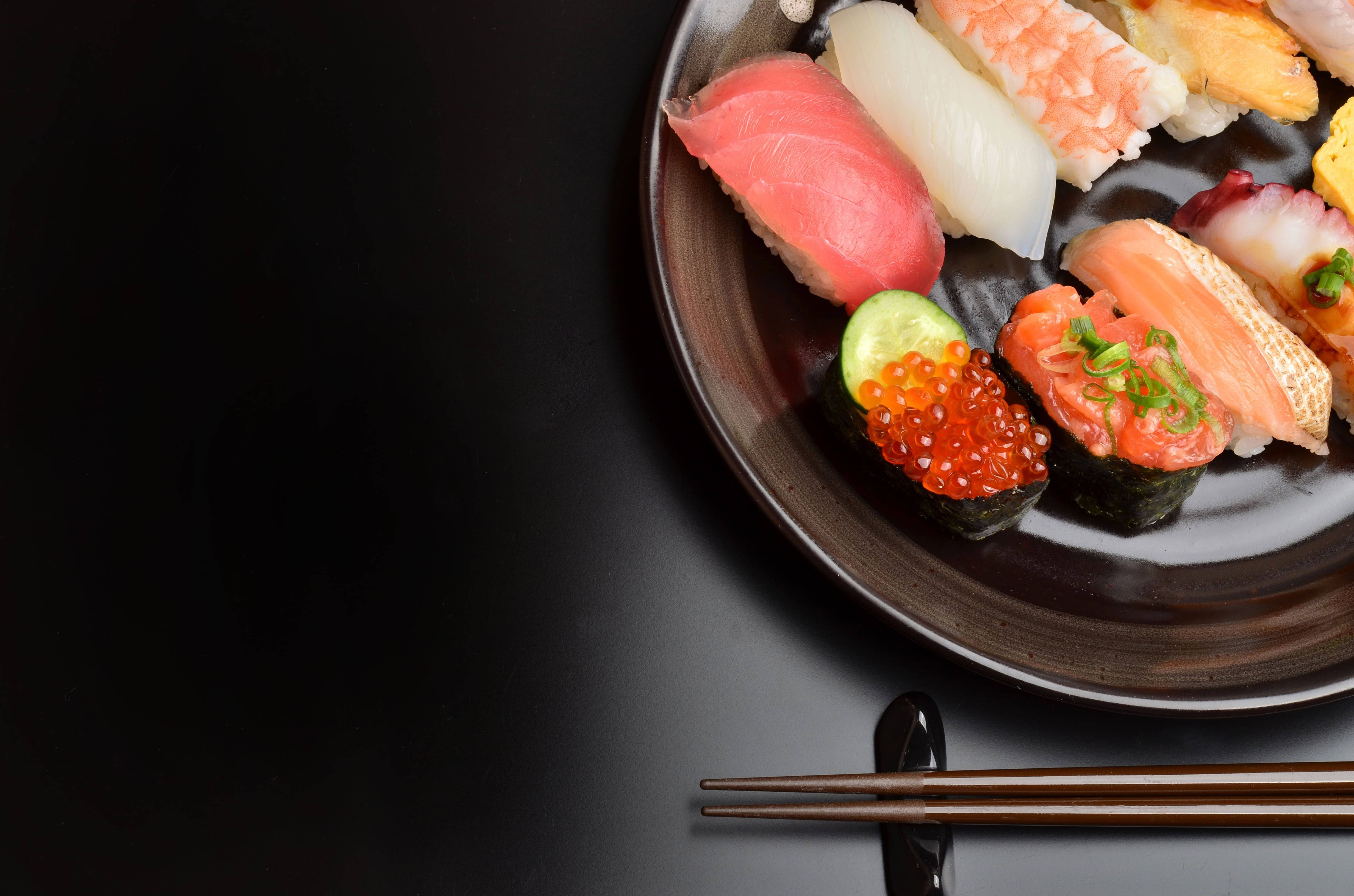 Sushi 桌布 壁紙 — 一個深色盤子上放著色彩鮮豔的壽司，包括鮪魚、鮭魚和魚卵，旁邊放著筷子，背景為黑色。
