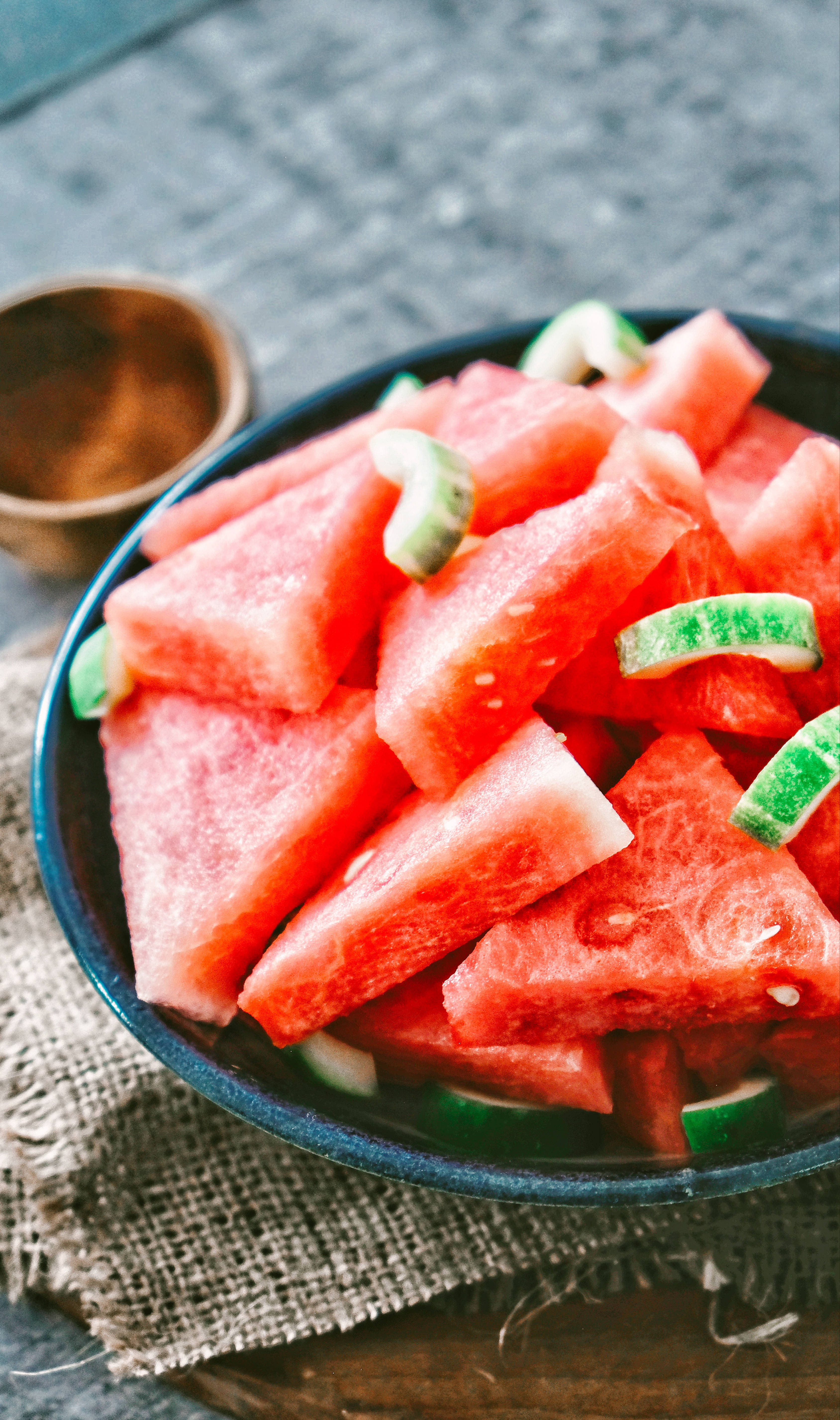 Watermelon 桌布 壁紙 — 一個深色碗裝滿多汁的紅色西瓜片和綠色瓜皮，放在木質表面上的質樸麻布上。