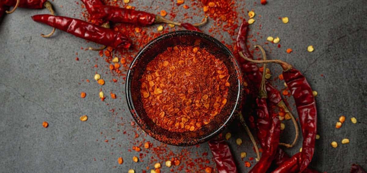 Red Chili Powder 桌布 壁紙 — 一個深色碗裝滿鮮紅色辣椒粉，周圍有乾紅辣椒和散落的種子，放在有紋理的灰色表面上。